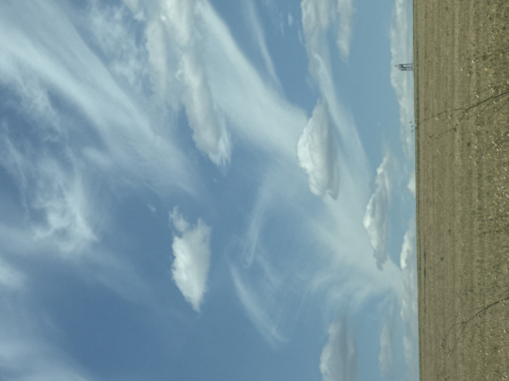 An expansive view of a vast, flat, dry field under a bright blue sky filled with wispy, streaky clouds. The field is a uniform, light brown color, suggesting a harvest or drought. In the far distance, a tall, slender metal tower structure is visible, standing on the horizon. The sky occupies approximately two-thirds of the frame, with the field filling the bottom third.