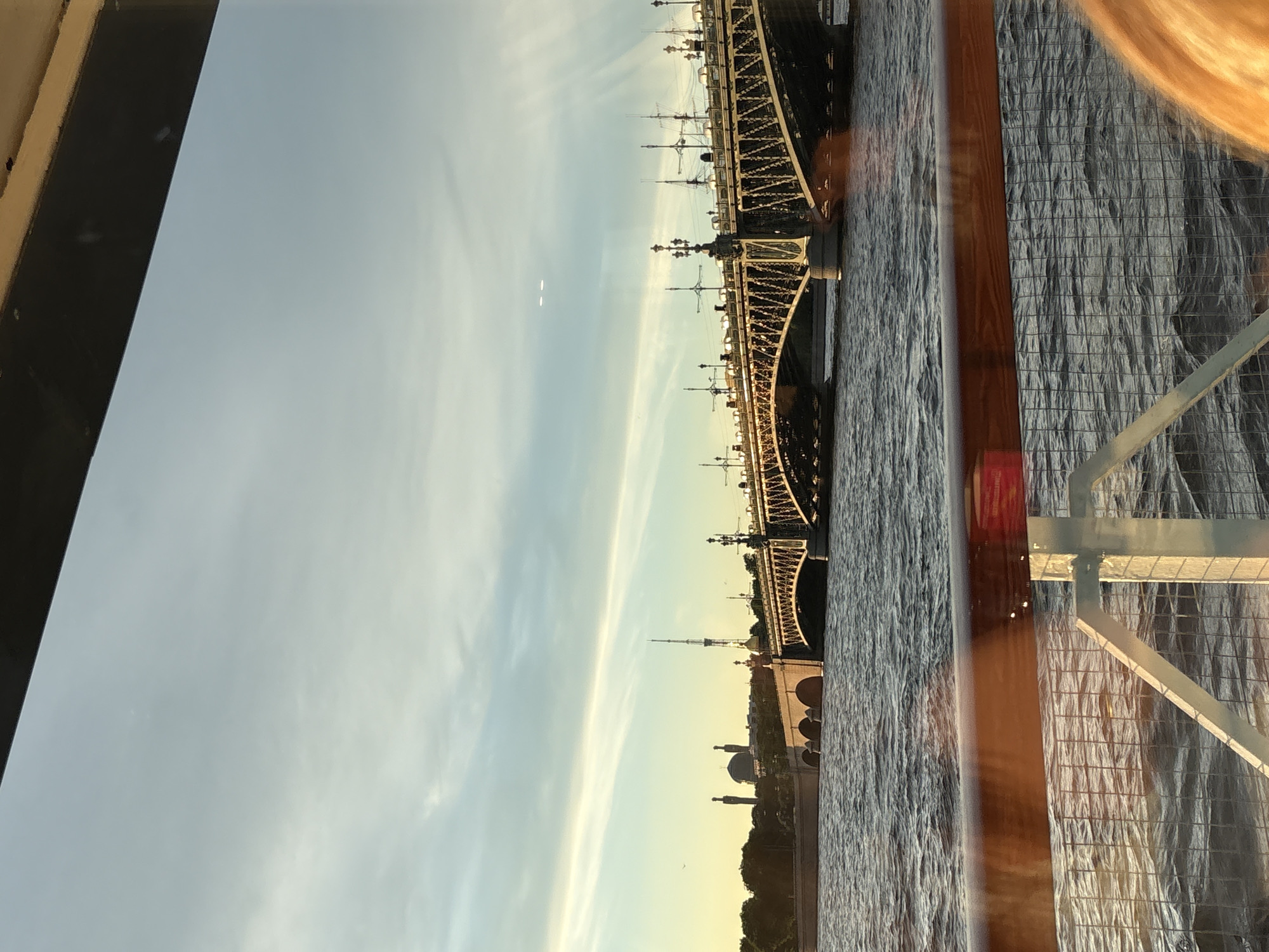 The image captures a view of the Troitsky Bridge in Saint Petersburg, Russia, at sunset. The bridge, an iconic landmark, spans across a body of water, likely the Neva River. The sky is a blend of warm yellow and orange near the horizon, transitioning to a pale blue with wispy clouds higher up, indicative of the golden hour.
In the background, the city skyline is visible, featuring distinct architectural elements. To the left, there are buildings with domes and minarets, suggesting the presence of religious structures. A tall, slender tower, possibly a communication or observation tower, stands prominently. The bridge itself is illuminated, with streetlights and other fixtures casting a warm glow. The water is choppy, reflecting the colors of the sky and the bridge.
In the foreground, a wooden railing and a metal mesh fence are visible, suggesting the vantage point is from a boat or a ferry. A portion of a person's blonde hair is visible on the right side of the frame, indicating that someone is present in the foreground. The overall impression is one of a serene, picturesque evening scene in the city.