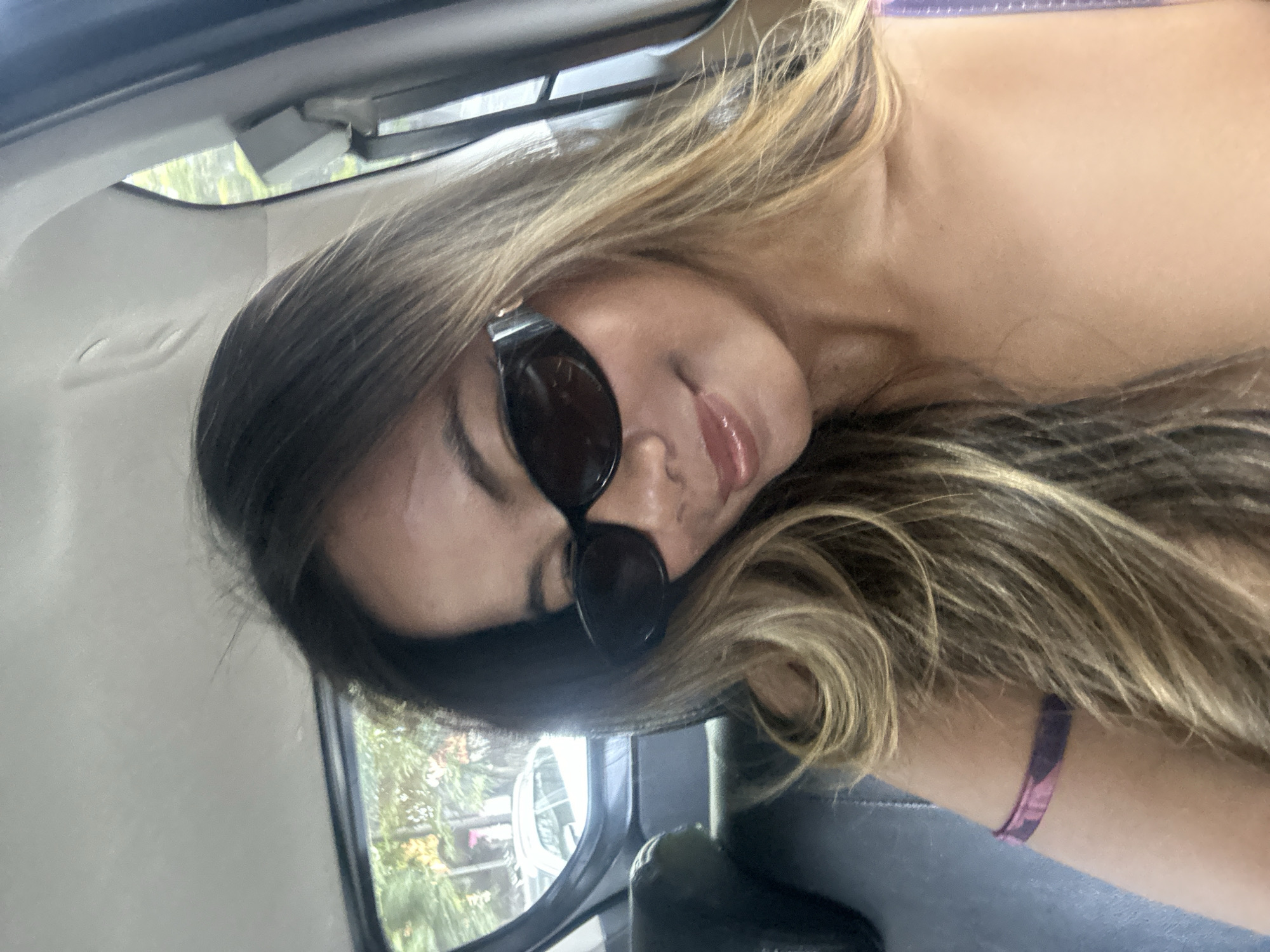 A young woman with long, wavy, light brown hair and sunglasses is inside a vehicle. She is wearing a pink and purple patterned strap, possibly part of a shirt or dress. The interior of the vehicle is visible, with a dark seat and a window showing a blurred outdoor scene of trees and a white car, indicative of an urban environment such as Denpasar, Indonesia. The woman is looking down and smiling slightly.