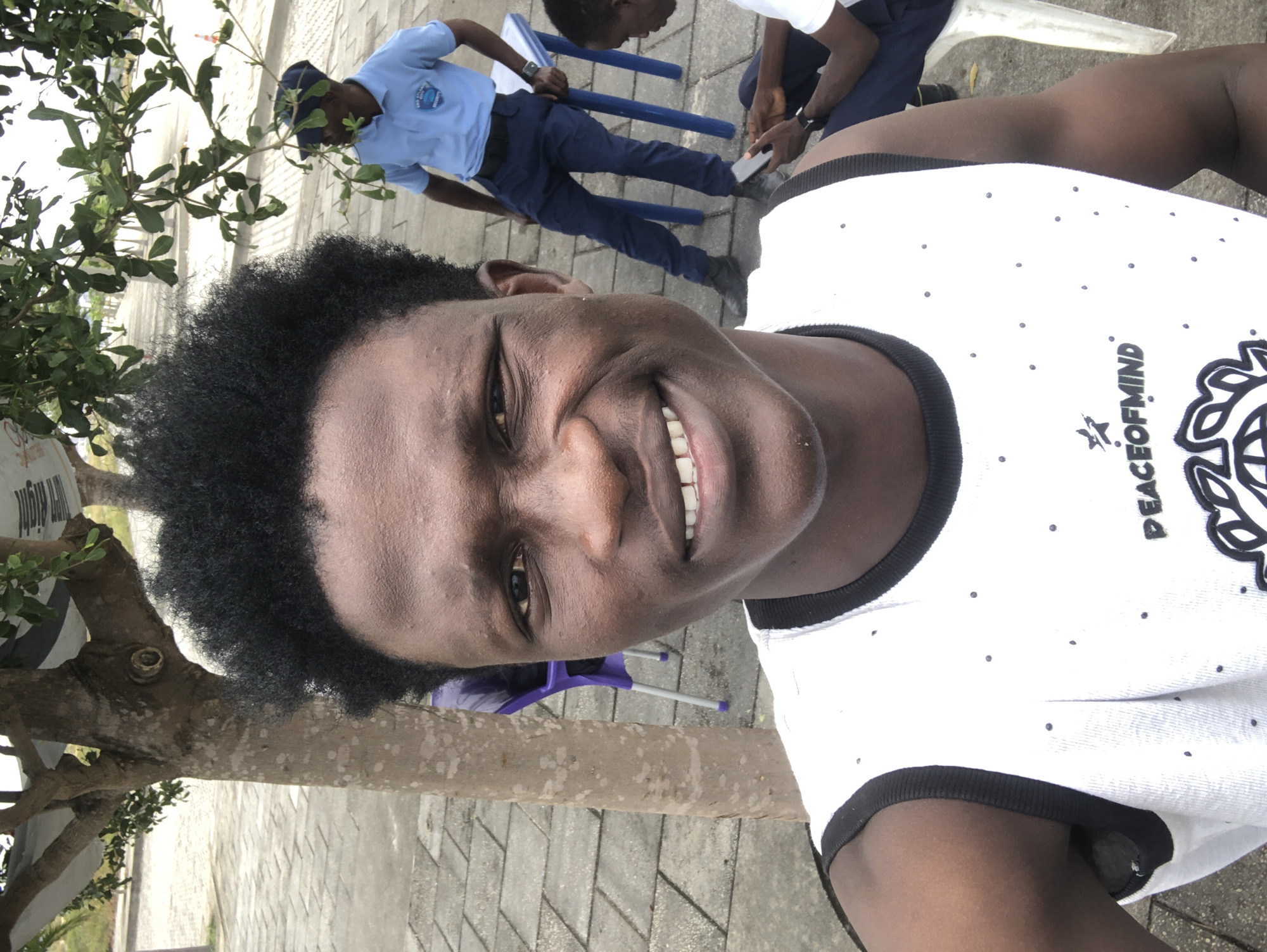 In Ikoyi, Nigeria, a young Black man with dark, curly hair and a wide smile takes a selfie. He is wearing a white sleeveless shirt with black trim and a pattern of small black dots. Behind him, a tree with light brown bark and green leaves stands to his left. In the background, a second Black man wearing a light blue polo shirt and dark blue pants, possibly security personnel, stands near a small blue table. Another person is partially visible sitting on a white chair to the right, looking at a mobile phone. The ground is paved with grey interlocking tiles, and a sidewalk is visible in the distance. The overall atmosphere appears to be outdoors, possibly a public space or park.