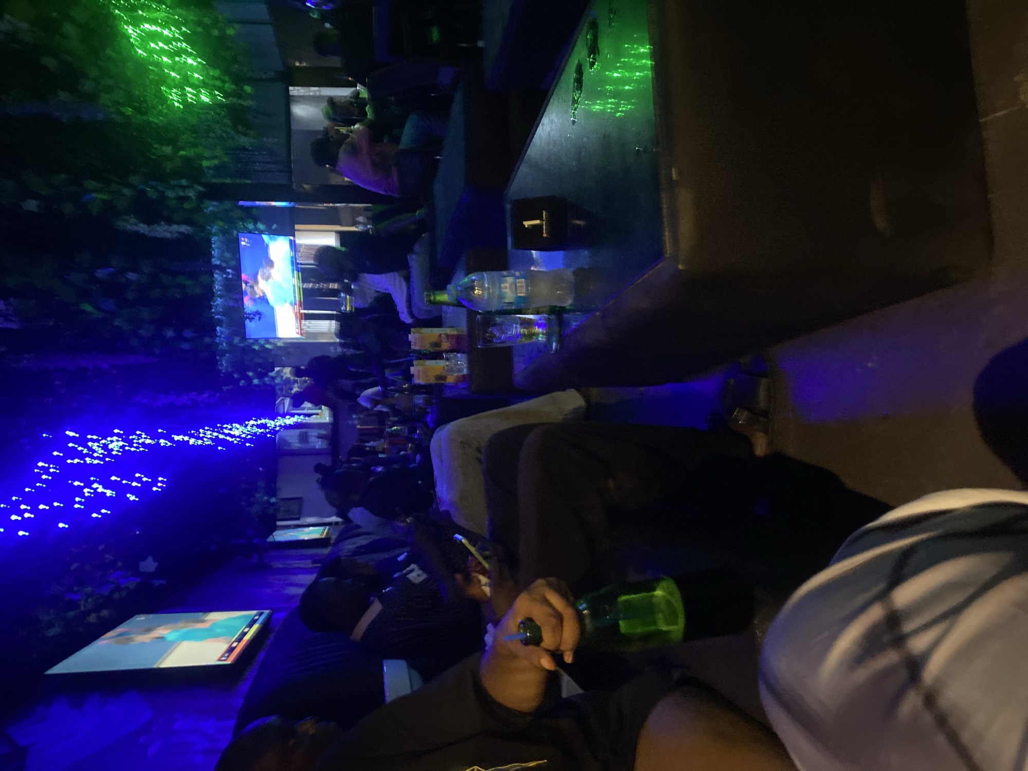 This media file captures a nighttime scene at an establishment, possibly a bar or lounge, in Abuja, Nigeria. The setting is dimly lit, with vibrant blue and green LED lights creating a festive and atmospheric ambiance. In the foreground, a person's hand holds a green glass bottle, suggesting they are either drinking or about to drink. Another person in the mid-ground is seen holding a similar bottle and appears to be in conversation. Several other individuals are seated around tables, some with drinks in front of them.
A prominent feature of the scene is a large television screen displaying what appears to be a sports broadcast or a similar visual program. The screen is illuminated, adding to the overall visual dynamic of the space. The decor includes what looks like artificial foliage, possibly with small white flowers, interspersed with the blue LED lights, creating a sense of an indoor garden or a nature-inspired theme. The presence of multiple screens and the gathering of people suggest a social event, perhaps watching a game or simply enjoying a night out. The overall mood appears relaxed and social, with the lighting contributing to a lively yet intimate atmosphere. The time of day is clearly night due to the artificial lighting being the primary source of illumination. There are no visible weather cues.