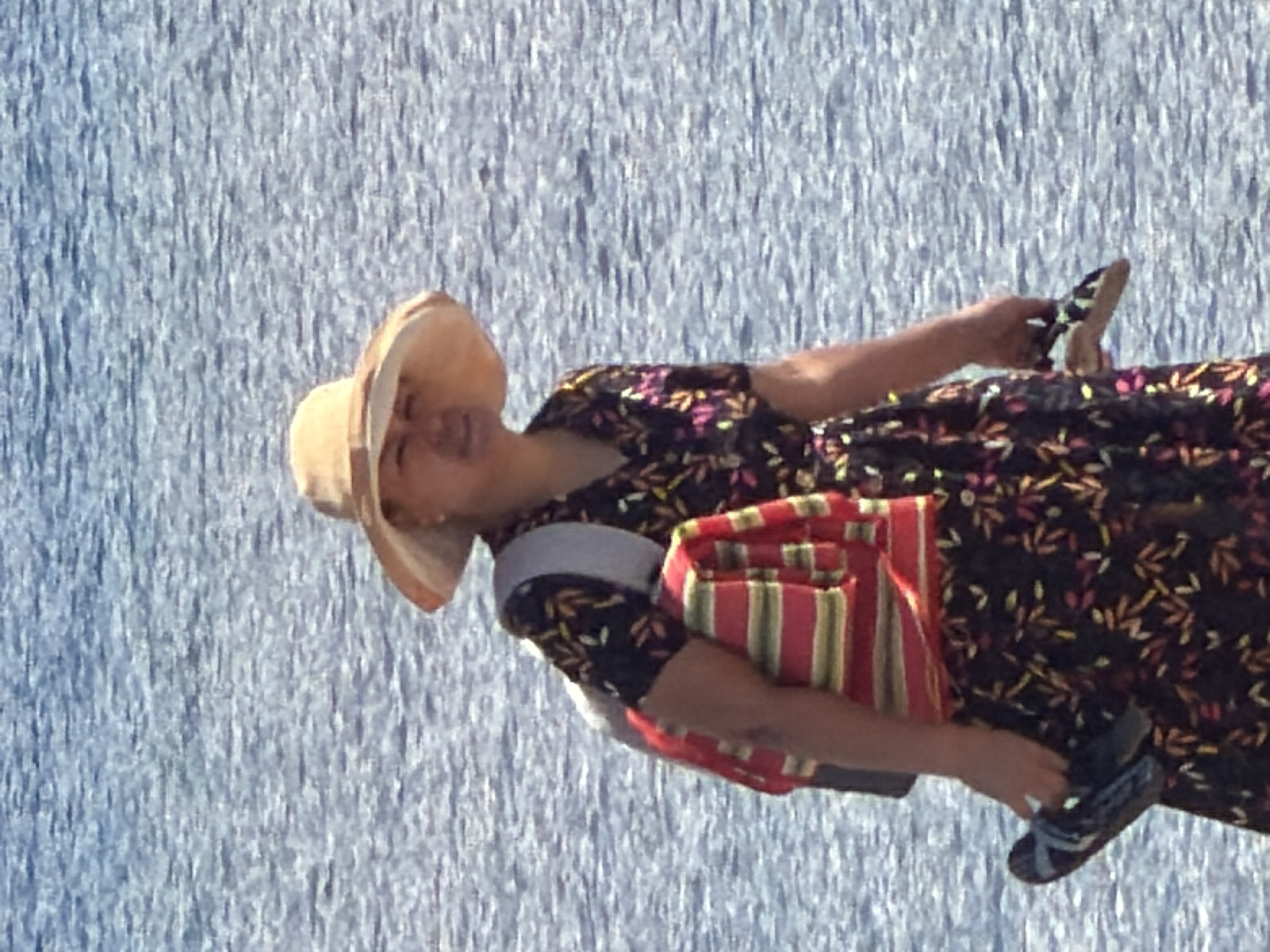 A person wearing a wide-brimmed hat and a floral dress stands in front of a body of water. The person is carrying a striped bag on their left shoulder and holding a pair of sandals in their right hand. The water behind the person is a blueish-grey color with ripples. The lighting suggests it is daytime.