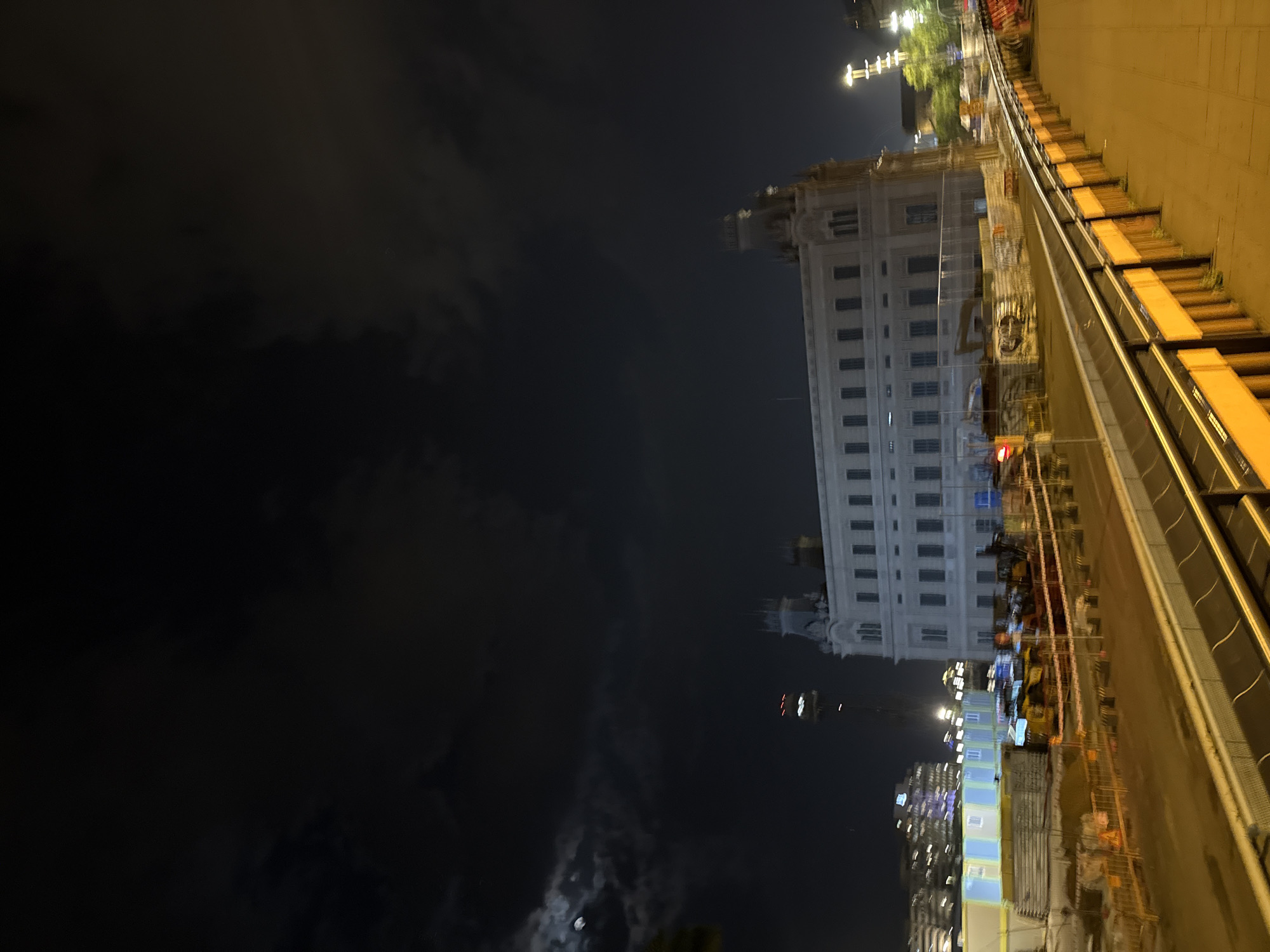 This nighttime image features a sideways view of Barcelona. A tall, white building with numerous windows stands beside a construction site along a walkway with yellow lighting. The building appears to be a government or institutional building. To the left of the building is a dark expanse which is likely the sea. The sky is black with clouds. In the distance are other buildings with lights.