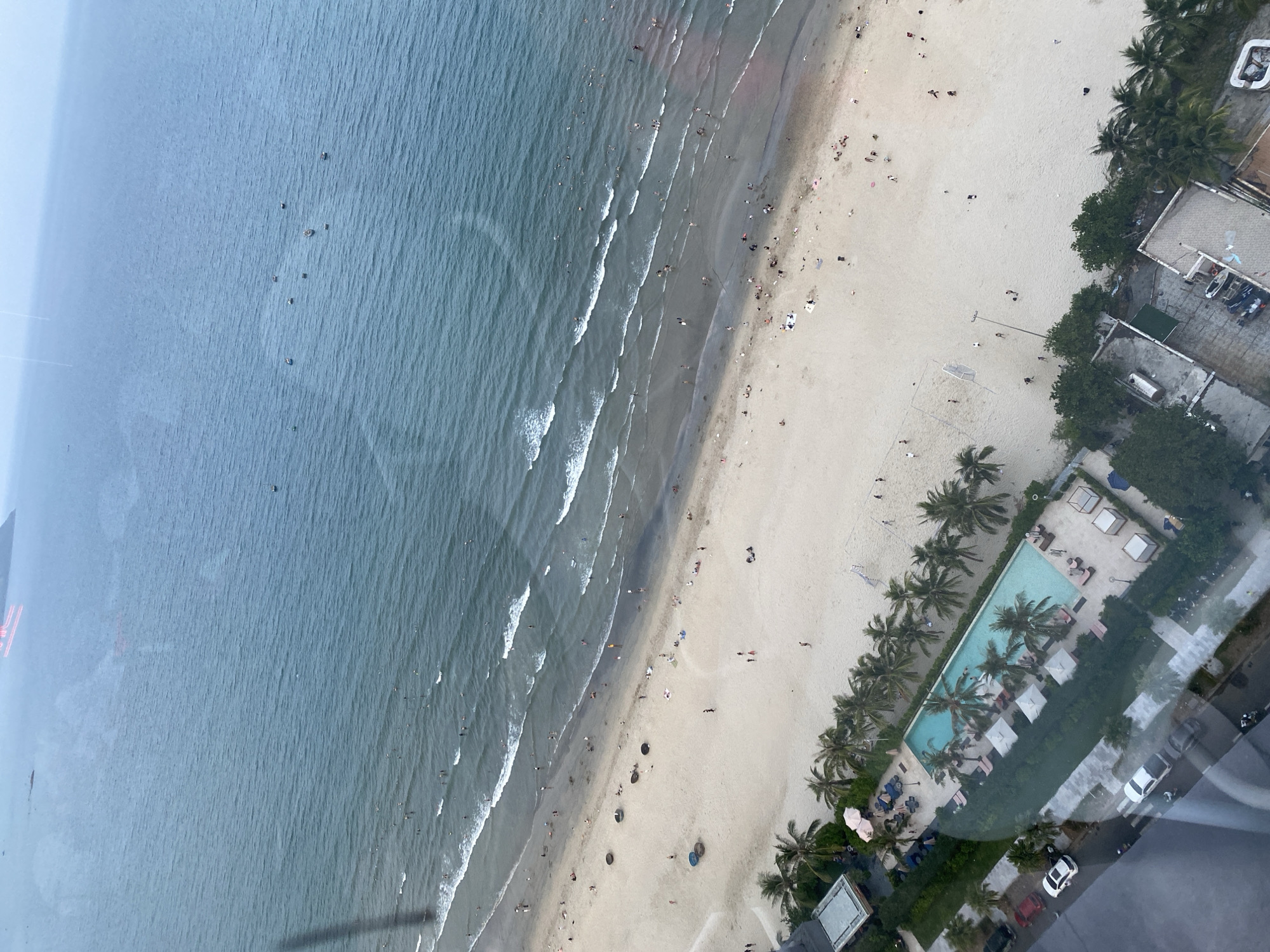This is an aerial view of a beach and resort area in Đà Nẵng, Vietnam. The ocean is a dominant element, with gentle waves lapping onto a sandy beach. Numerous people are visible scattered across the beach, some swimming in the ocean and others relaxing on the sand.
Adjacent to the beach is a resort area with a prominent swimming pool, surrounded by lounge chairs and umbrellas. Palm trees are interspersed throughout the resort, adding to the tropical atmosphere. Several buildings, likely part of the resort or nearby hotels, are visible, along with a road where cars are parked. In the distance, more boats are dotted across the ocean. The overall scene depicts a busy beach and recreational area on a slightly overcast day.