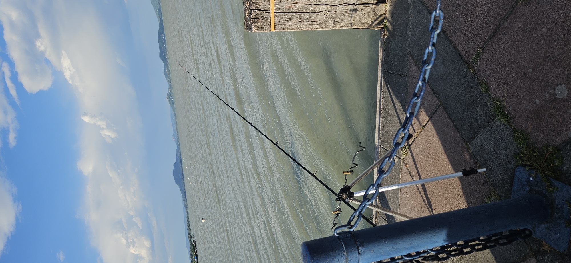 The image captures a serene lakeside scene with a fishing rod set up on a tripod, poised over the water. The fishing rod is black with a white fishing line extending into the murky green water. A blue metal post with a chain attached stands to the left of the tripod. To the right, a weathered wooden post with a yellow band serves as a boundary marker. The foreground consists of a paved walkway made of interlocking gray and reddish-brown stones, with patches of grass growing between them. In the background, the calm lake stretches towards a distant, hazy shoreline with rolling hills and mountains under a bright blue sky dotted with fluffy white clouds. The overall atmosphere is peaceful and quiet, suggesting a solitary fishing activity. The location is identified as Balatongyörök, Hungary, a resort town situated on the northern shore of Lake Balaton.