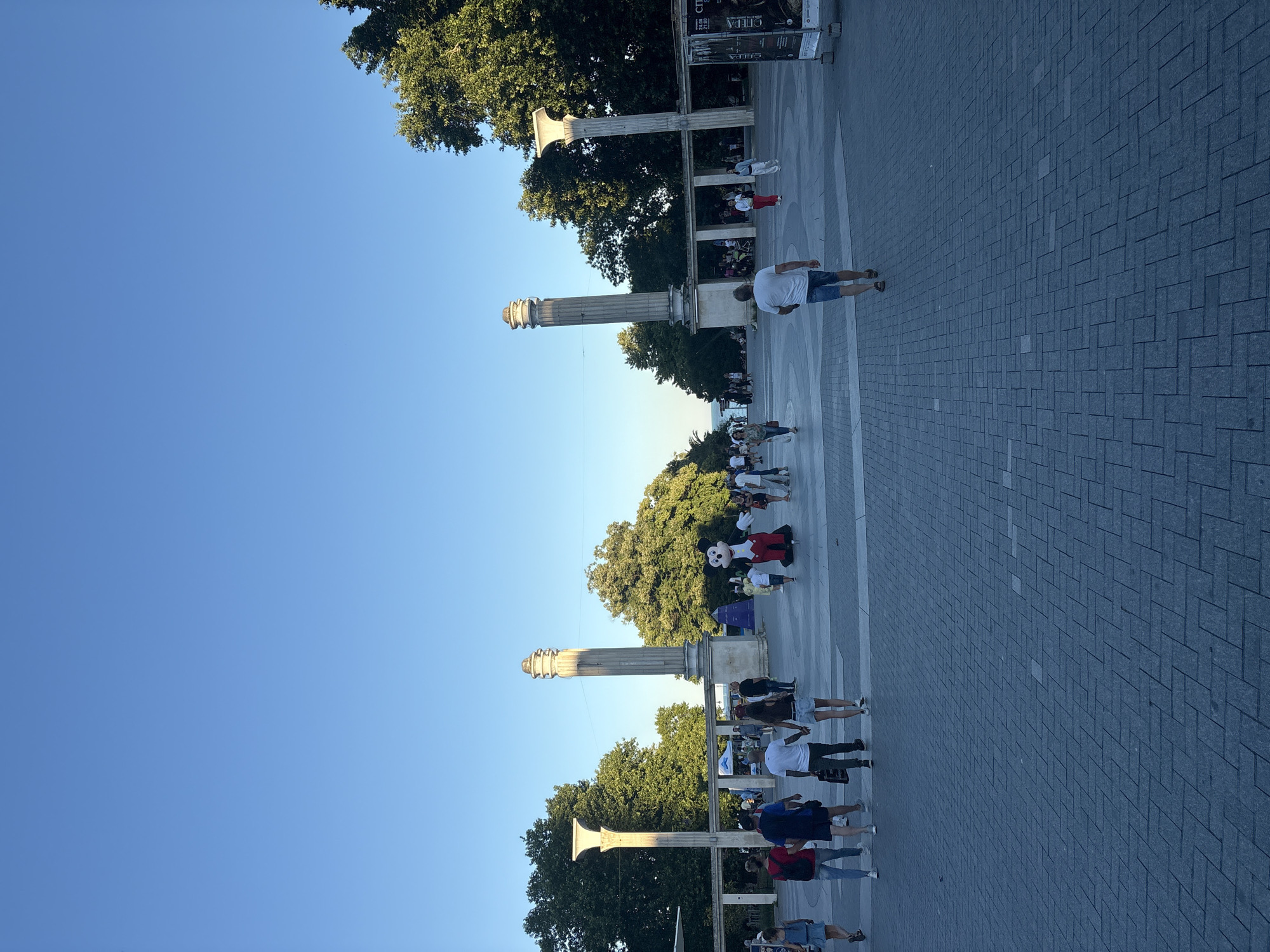 An outdoor, low-angle, medium shot captures a public square paved with grey interlocking pavers under a clear blue sky. The scene is illuminated by the warm, low light of what appears to be late afternoon or early evening. On the right side of the frame, a large, inflatable Mickey Mouse character stands in the middle of the square, facing slightly to the left and appearing to interact with the crowd.
A diverse group of people are scattered throughout the square, engaged in various activities. Some are walking, some are standing, and a few appear to be observing the Mickey Mouse character. A couple holding hands walks away from the camera in the foreground, while a man in a white t-shirt and blue shorts walks towards the camera. In the background, a line of people is visible near a series of large, classical-style columns and arches, suggesting a significant architectural feature or entrance. Lush green trees frame the scene on both sides and behind the columns, indicating a park-like or landscaped urban setting. A tall, slender, fluted column with a decorative top stands prominently on the left, balanced by another similar column on the right, with a covered walkway or arcade connecting them. On the far right, a tall, narrow billboard with text and images is visible, suggesting an event or advertisement. The overall atmosphere is one of leisure and public gathering.