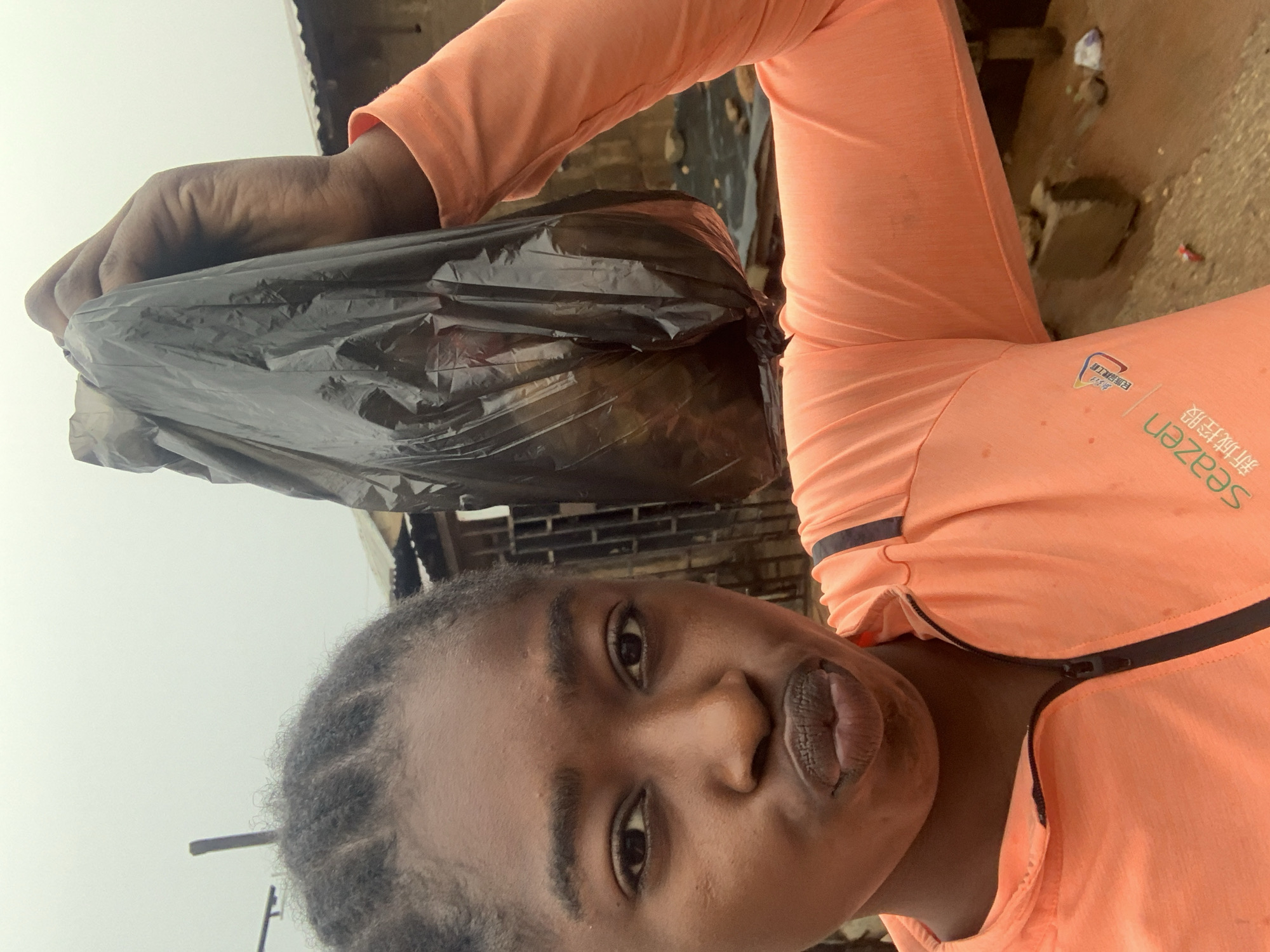 A person is holding a black plastic garbage bag, apparently near the New Stadium in Nigeria. The person is wearing a long-sleeved coral-colored shirt with a zipper. In the background are buildings of brick and corrugated metal, and scattered debris on the ground. The overall impression is one of poverty or a developing area.