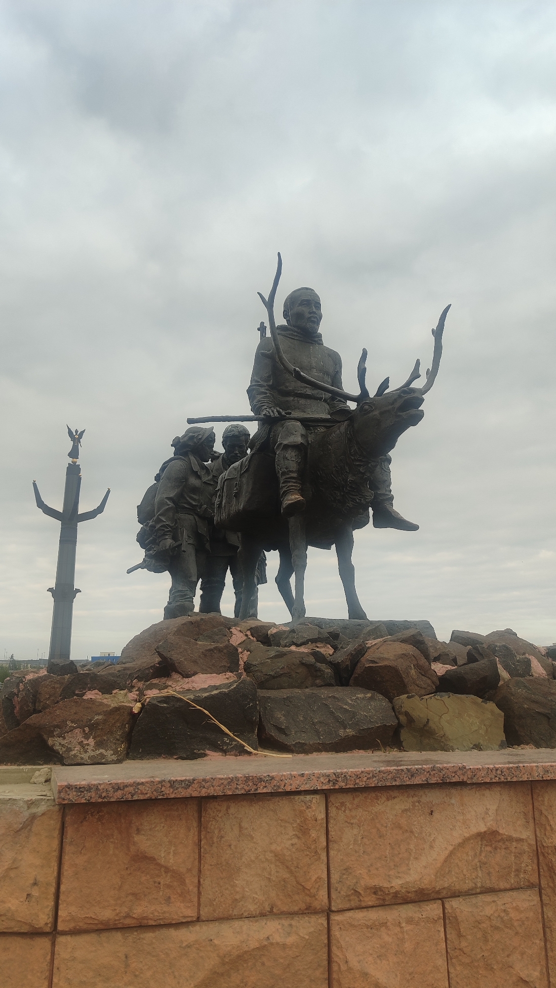 A bronze statue depicts three figures and a reindeer. The central figure, a man, sits astride the reindeer, holding a bow and arrow. Two other figures, a man and a woman, stand beside the reindeer, looking towards the central figure. The statue is mounted on a rocky base. In the background, a tall, dark monument with a winged figure at the top stands against a cloudy sky. The scene is located in the city of Mirny, Russia.