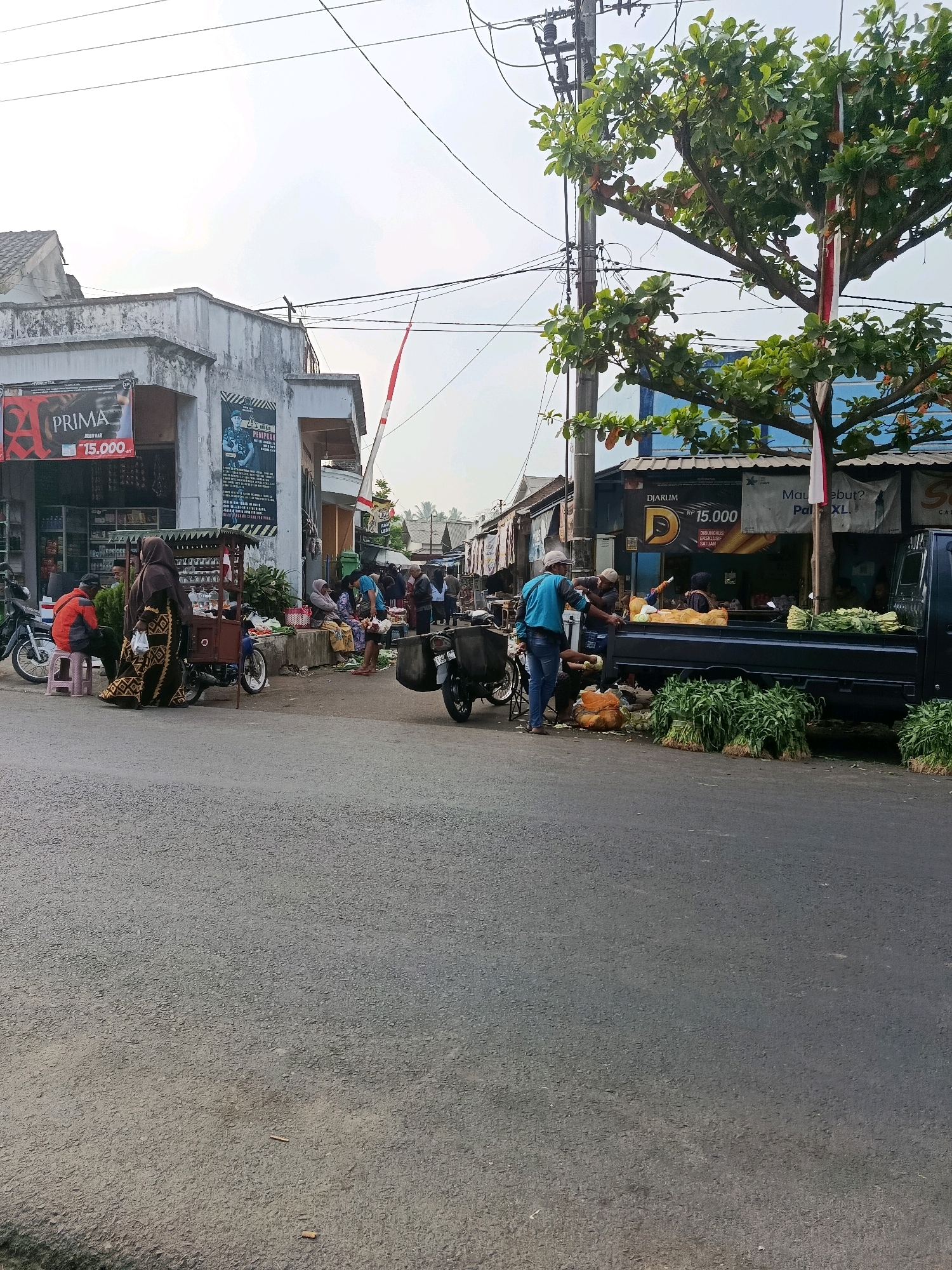 This is a photograph taken from a street-level perspective in Tajinan, Indonesia. The scene depicts a bustling roadside market with a variety of stalls and vendors. In the foreground, the asphalt road is visible, with a slight incline.
To the left, a building with a faded facade houses several shops. One storefront displays a large red and white banner with the word "PRIMA" prominently featured, along with "Rp 15.000". Further down the street, a smaller building displays a poster of a person in uniform.
Several individuals are engaged in market activities. A woman in a patterned abaya stands next to a mobile cart, possibly selling goods. A man in an orange jacket sits on a pink stool, and another person is seated nearby. On the right side of the image, a dark pickup truck is parked, laden with fresh produce, including large bunches of green vegetables. Two men are interacting near the truck, one wearing a blue shirt and the other a cap. More produce is arranged on the ground around the truck.
Other elements of the environment include a utility pole with numerous wires extending overhead, a tree with green leaves and some reddish-brown fruits, and a red and white striped barrier, possibly associated with a level crossing or a local landmark. The sky is overcast, suggesting a cloudy day. The overall atmosphere is that of a typical local market on a street.