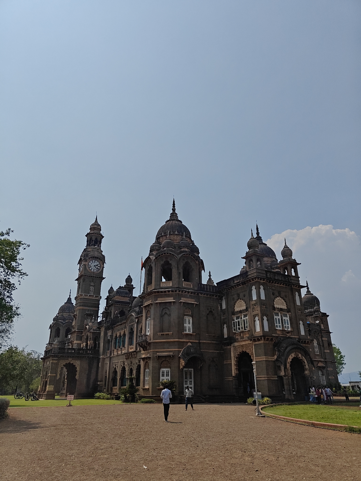 An exterior, low-angle, wide shot depicts the New Palace (Shri Chhatrapati Shahu Maharaj Palace) in Kolhapur, Maharashtra, India, under a clear, light blue sky with minimal cloud cover on the right. The palace is a large, multi-domed, multi-spired structure constructed of dark grey or brown stone, featuring Indo-Saracenic architecture. A prominent clock tower, adorned with a circular clock face, stands on the left side of the building. A red flag is visible on a spire to the right of the clock tower. The palace façade displays numerous arched windows, decorative carvings, and balconies.
In the foreground, a wide, reddish-brown unpaved pathway curves towards the palace. Patches of green lawn are visible on either side of the pathway closer to the building. On the far left lawn, two historical cannons are positioned. A small yellow sign with unreadable red text stands on the lawn to the left. A white lamppost or pole is present on the right edge of the pathway.
Multiple individuals are present. One male, viewed from behind, wearing a light-colored shirt and dark trousers, walks centrally on the pathway towards the palace. Another male in a plaid shirt is partially visible near an entrance on the left side of the palace. A group of approximately 5-7 individuals is gathered near an entrance on the right side of the palace, appearing to enter or exit. Green foliage from trees is visible on the far left and in the distant background.