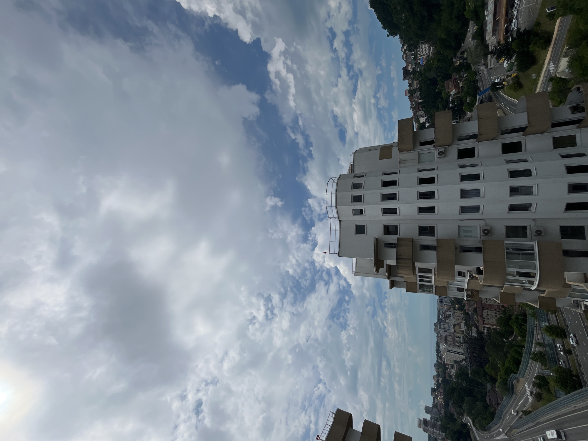 This image shows a unique, stepped white building with numerous windows and balconies, likely an apartment complex, situated in Sochi, Russia. The building's unusual architecture is prominent in the foreground, with the curved balconies projecting outwards. In the background, a cityscape with other buildings, a multi-lane road with light traffic, and lush green hills covered in trees can be seen. The sky is filled with a mix of white and gray clouds against a blue sky. The scene captures a part of the urban environment of Sochi, with the distinct building serving as a focal point.