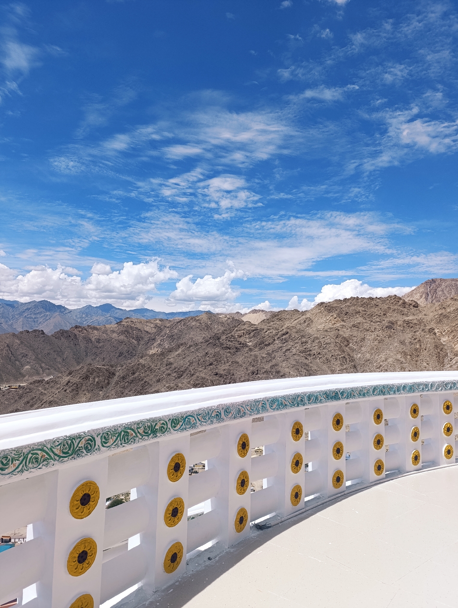 The image shows a white railing decorated with green and yellow floral patterns and circular motifs. Beyond the railing, a vast, arid mountain landscape stretches towards the horizon under a bright blue sky with scattered white clouds. The mountains are rugged and appear to be sparsely vegetated. The scene is likely a viewpoint or a rooftop terrace in Leh, India, offering a panoramic view of the surrounding mountainous terrain. There are no people visible in the image.