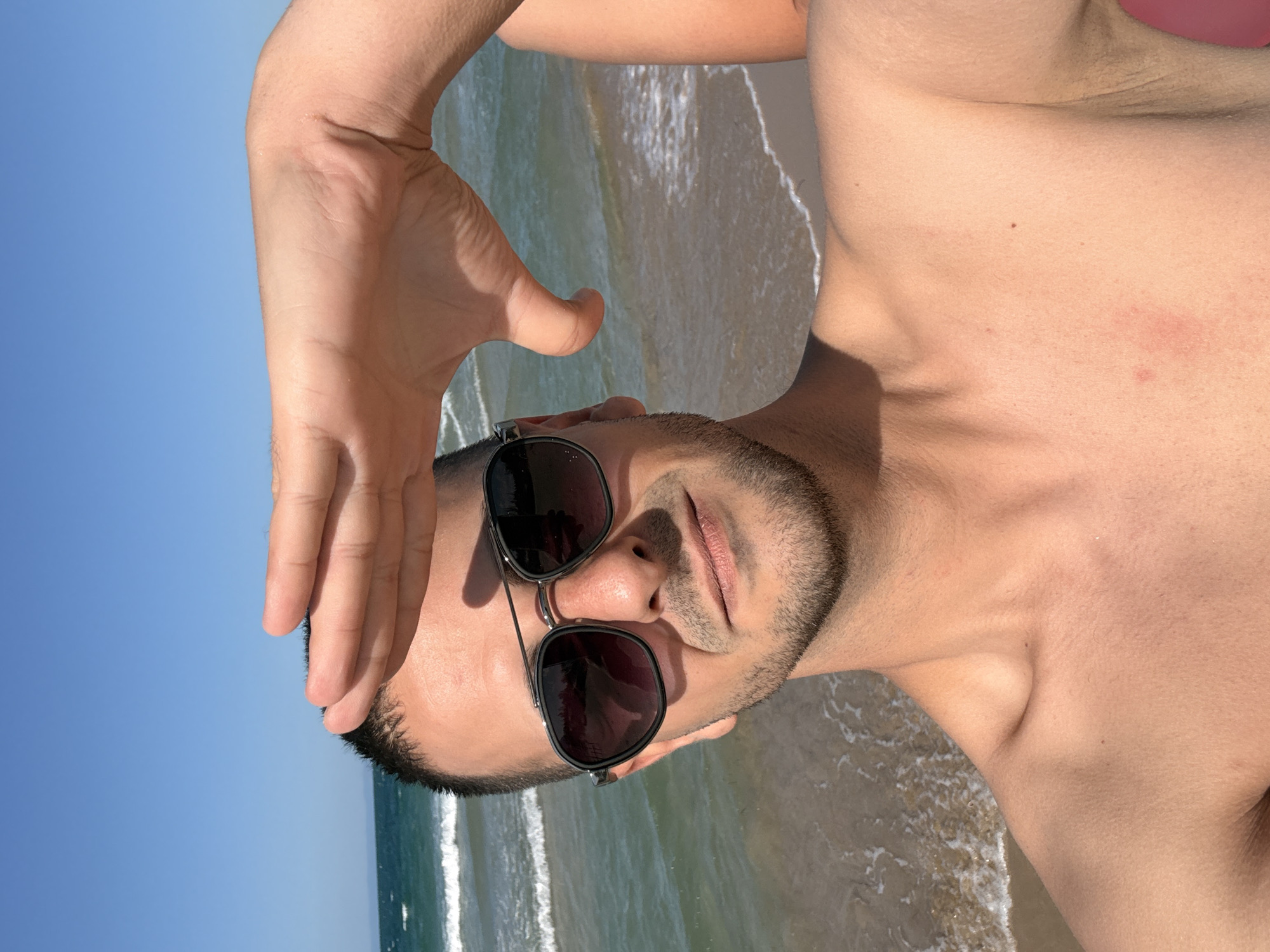 A man with short dark hair, sunglasses, and a light beard is taking a selfie on a beach in Sozopol, Bulgaria. He is shirtless and holding his hand up to shield his eyes from the sun. The sea and sky are visible in the background, with gentle waves breaking on the shore. The overall lighting suggests a sunny day.