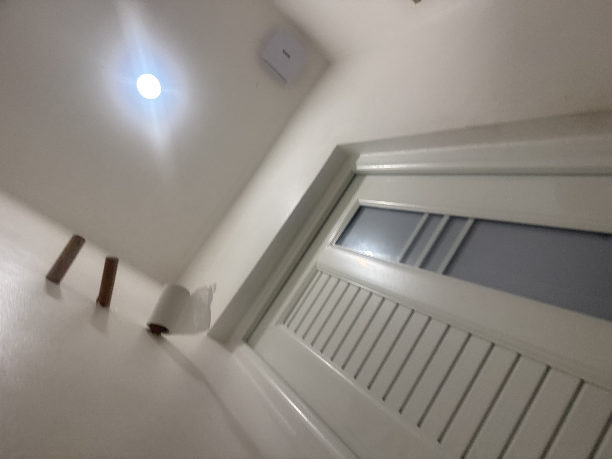 The image depicts a low-angle shot of a white door with louvered panels and a rectangular glass insert. Above the door, on a white ceiling, are two small, brown wooden pegs mounted horizontally, with a roll of toilet paper suspended from the lower peg, partially unspooled. A bright, circular light fixture is visible on the ceiling to the right, casting a glare. A small, white square ventilation or smoke detector is mounted on the wall in the upper right corner. The scene appears to be an interior, possibly a bathroom or hallway, in Kuta Selatan, Indonesia.
