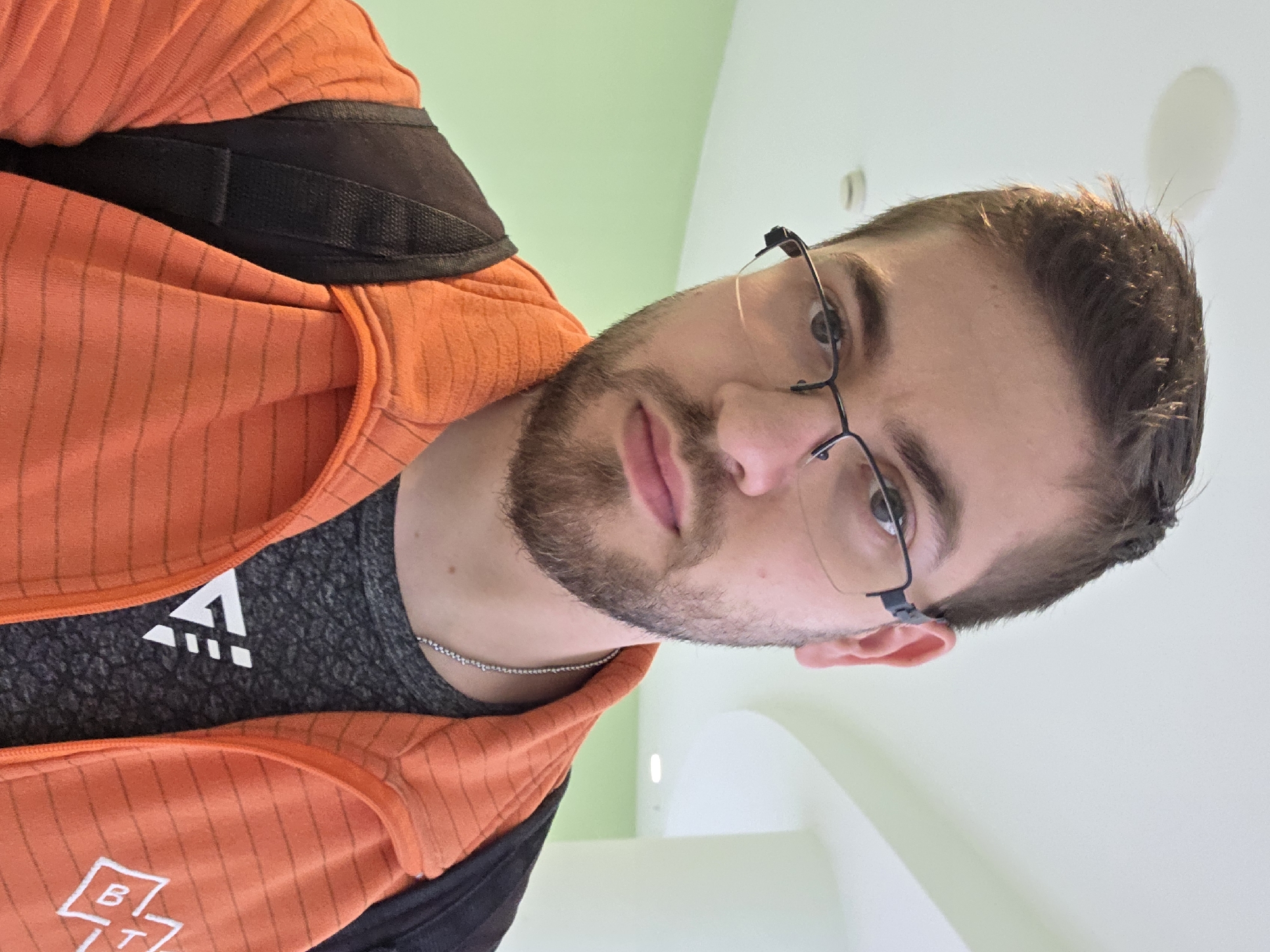 A person with fair skin, brown hair, and a beard is visible in the image. They are wearing glasses with thin black frames. The individual is wearing a gray t-shirt under an orange and black jacket. A thin silver chain is visible around their neck. The background is a blend of pale green and white. There is no specific location in Plovdiv, Bulgaria, identifiable in the provided image.