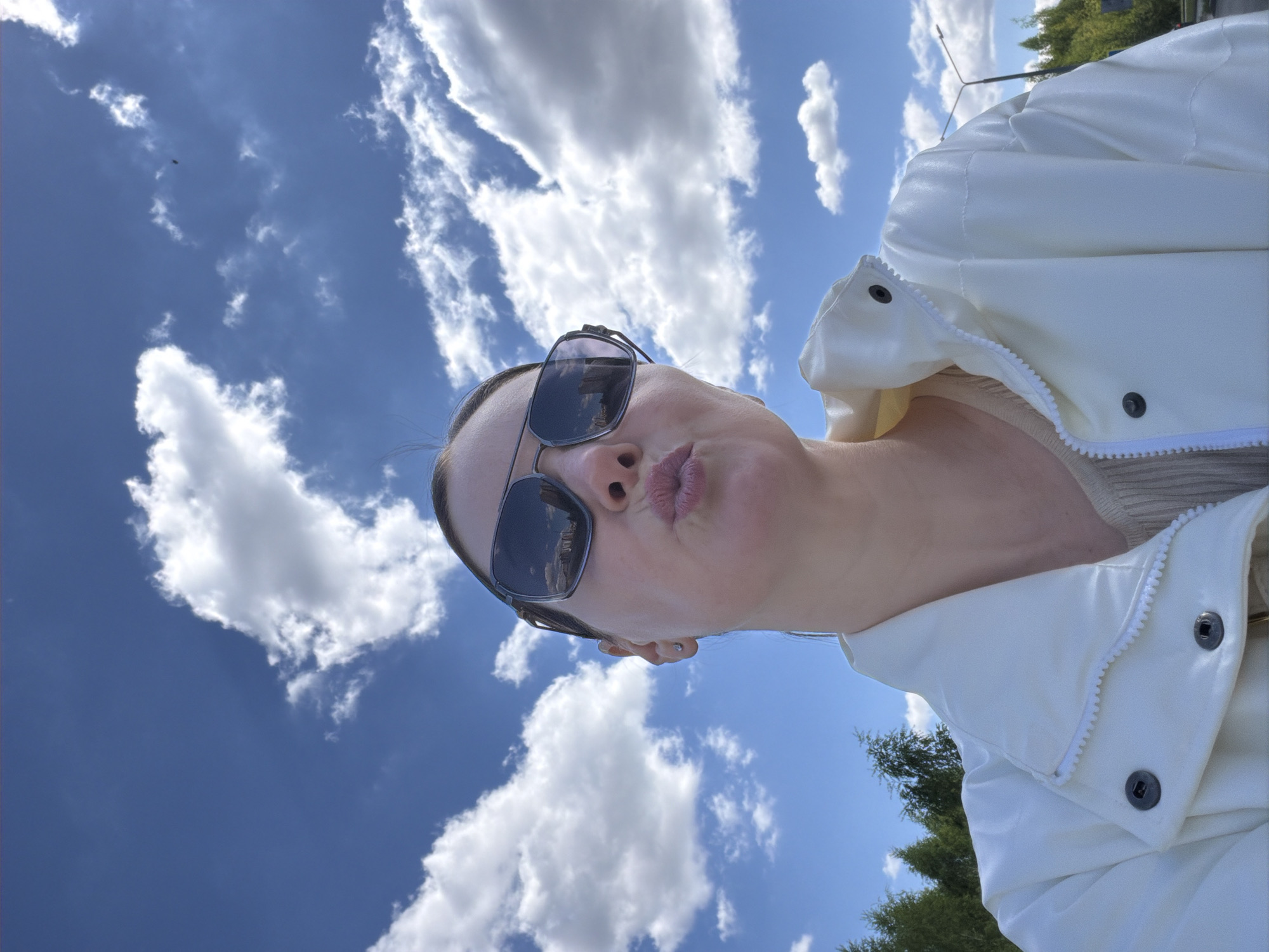 A rotated medium shot depicts a woman wearing sunglasses and a white jacket against a backdrop of a blue sky with puffy white clouds. The woman is facing the camera with her lips pursed. Part of a tree is visible in the lower right corner. The location is in the city of Unknown, Unknown.