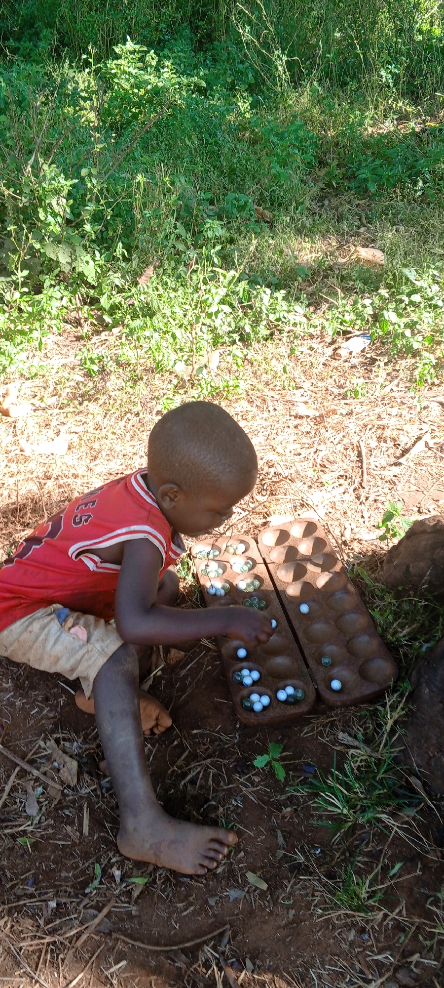 In an outdoor setting within Ngalula, Democratic Republic of the Congo, a young boy is kneeling on the ground, engrossed in playing Mancala. The board game, carved from dark wood, rests directly on the bare earth amidst scattered leaves and sparse vegetation. The board's shallow depressions are filled with clear and white marbles. The boy, wearing a red jersey with white trim and light-colored shorts, is barefoot and focused on manipulating the marbles within the game. The background consists of grassy terrain and dappled sunlight filters through the trees, illuminating the scene.