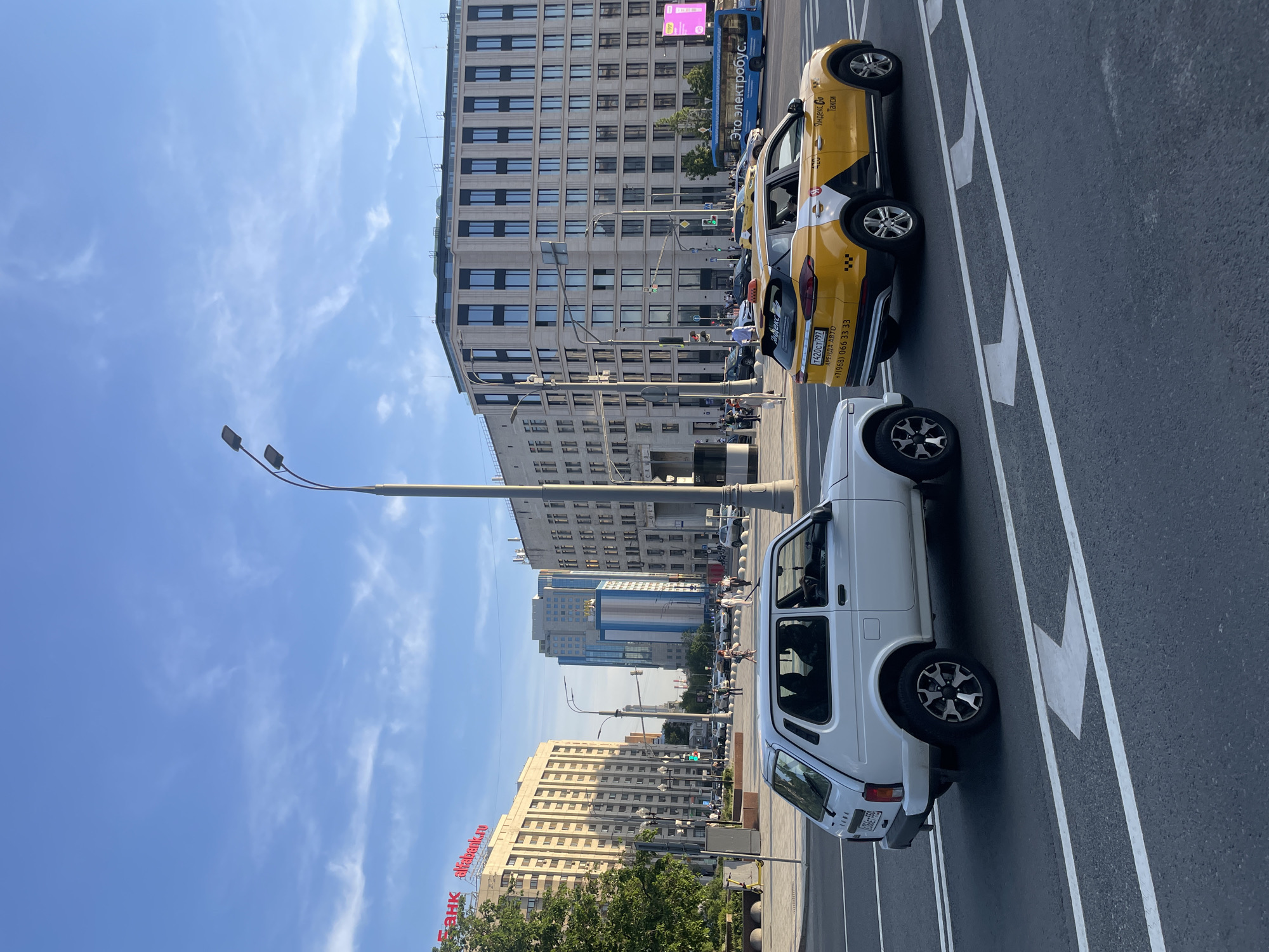 The image shows a street scene in Moscow, Russia on a sunny day. There are two cars parked along the street with painted directional arrows. The first car is a white Lada Niva, and the second is a yellow taxi with black and white accents, bearing the text "Яндекс Такси". There are several pedestrians in the background. Buildings of varying heights and architectural styles line the street. Signage for "Альфа-Банк" is visible on the building on the left. A street lamp is also visible. A blue "Электрический автобус" (Electric Bus) bus is visible behind the taxi cab. The license plate of the taxi cab is visible: T420CT797.