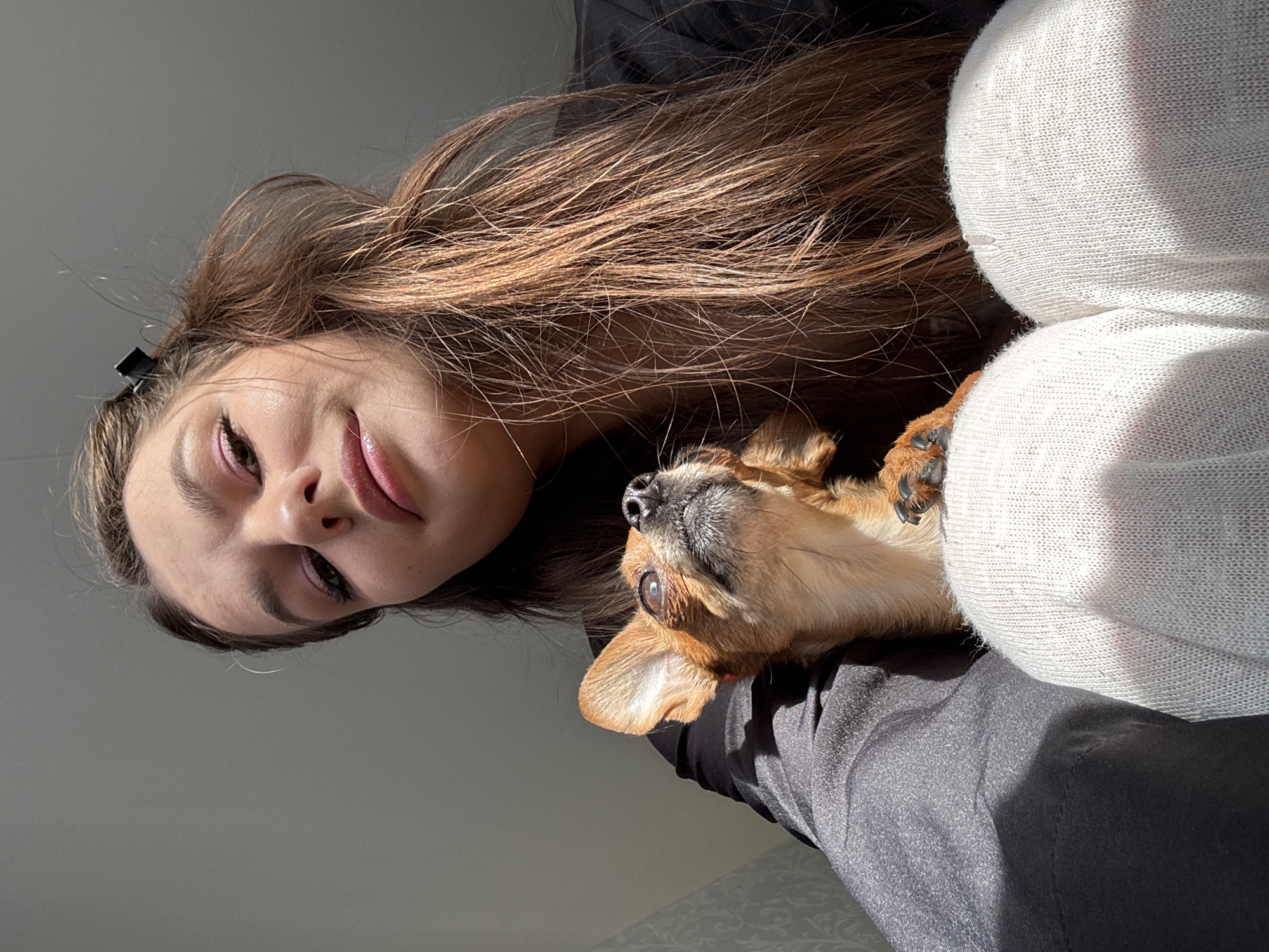 A low-angle, close-up shot depicts a woman with long brown hair and a small tan chihuahua. The woman has fair skin, pink eye shadow, and glossy lips. A black hair clip is visible in her hair. The chihuahua is nestled between the woman and a person wearing a white knitted garment. The dog is looking upwards. The woman is wearing a dark gray shirt. The location is likely indoors with sunlight entering the frame. The city of Chaikovsky, Russia, cannot be verified from the visual information provided.