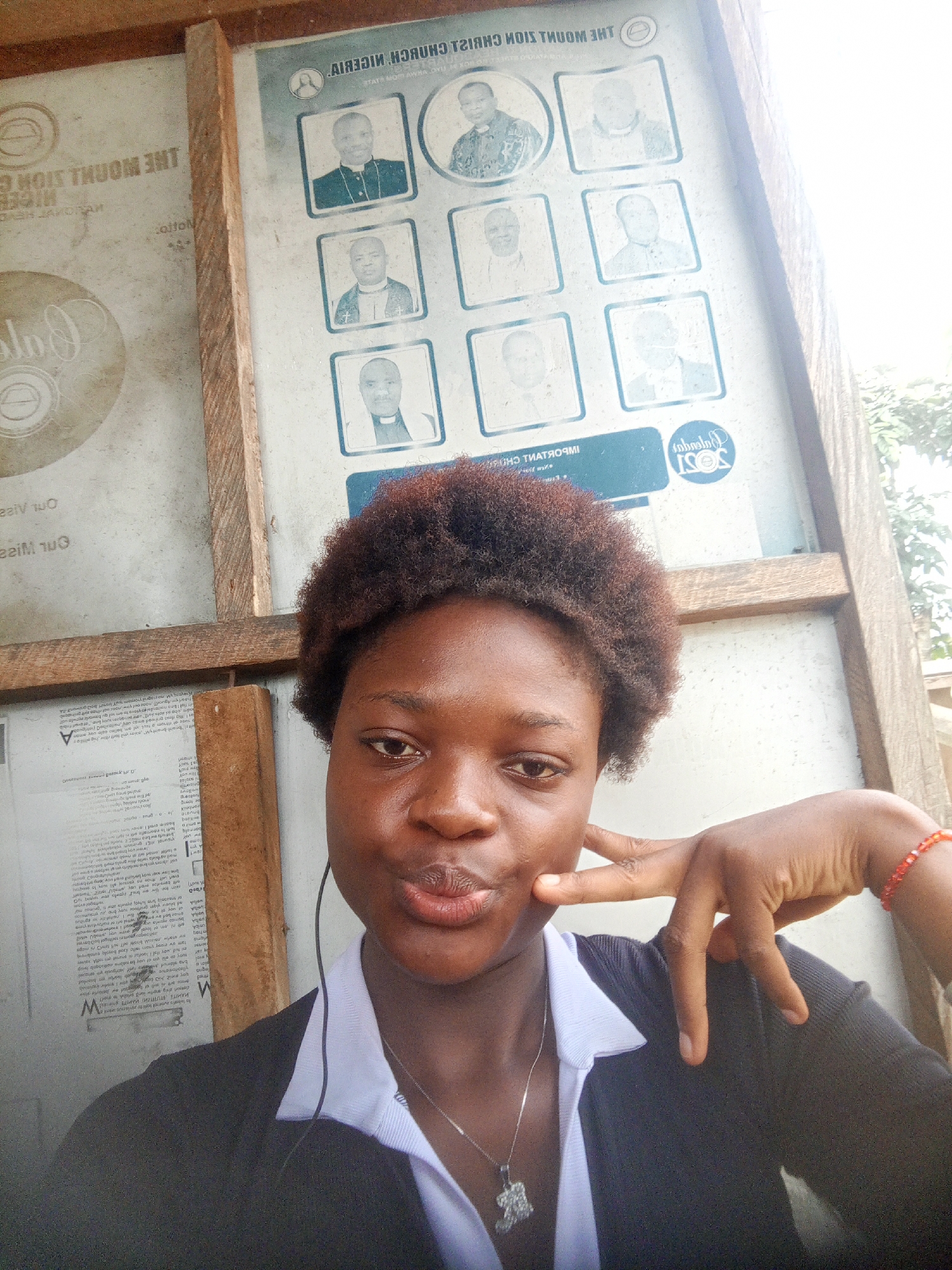 A young woman poses for a selfie in front of a bulletin board. The board is in a wooden frame, with several posters attached. One poster shows portraits of religious figures. A logo for "SOSI 2021" is visible on another poster. The woman has dark skin and short, curly hair. She is wearing a black cardigan over a white collared shirt, and a necklace with a cross pendant. She is making a peace sign with her fingers. A red beaded bracelet is on her wrist. The name "THE MOUNT ZION CHRISTIAN CHURCH NIGERIA" is visible on one of the posters. The environment appears to be outdoors, possibly near the city of Mbribit, Nigeria, based on the name of the church.