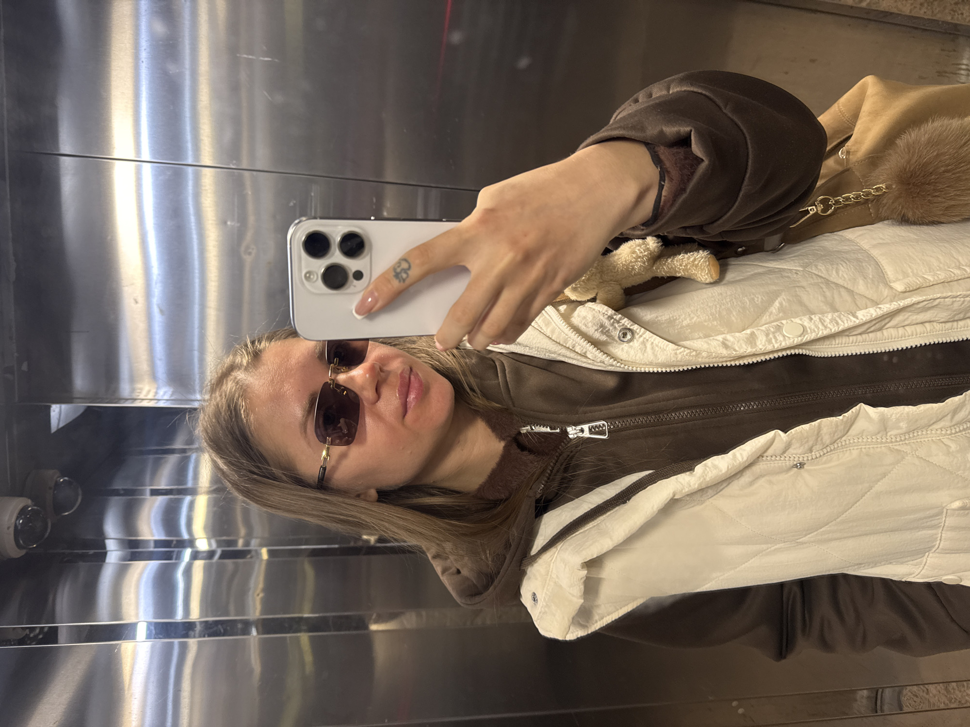 A woman with shoulder-length blonde hair, wearing brown-tinted sunglasses, is taking a selfie in an elevator. She is holding a white smartphone with multiple camera lenses. Her right hand, which is holding the phone, has a small tattoo on one of her fingers. She is wearing a brown jacket with a white quilted vest over it. A beige purse with a fur pom-pom attached to the strap is visible. The elevator has a metallic, mirrored interior. The location is likely within the city of Batumi, Georgia, though there are no explicit location-based clues in the frame of the photograph.