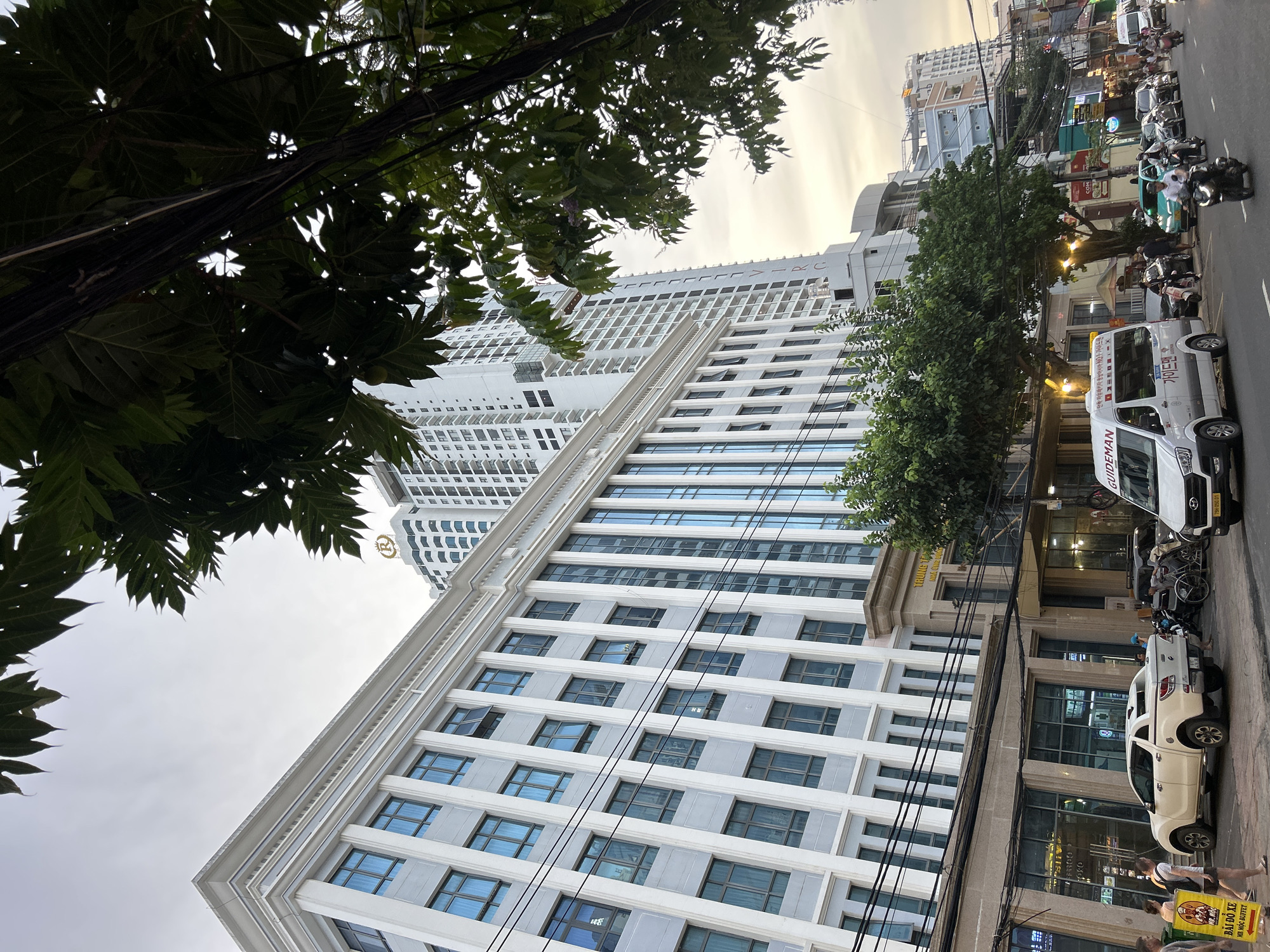 A bustling urban street scene is captured, characterized by a mix of modern and traditional architecture under an overcast or late afternoon sky. The image features a prominent tall white high-rise building with a distinctive pattern of windows and a large "H" or similar logo near its peak, alongside a shorter, classical-style building with multiple stories and blue-tinted windows. Numerous black power lines crisscross the upper part of the frame, partially obscured by lush green tree foliage.
The street level is active with vehicles and people. A white "GUIDEMAN" van with Korean text ("단독 자유패키자 동남아시아 NO.1 가이드맨" - Independent free package Southeast Asia No.1 Guideman) is parked, and a white pickup truck is also visible. Many motorbikes, both parked and in motion, populate the road, indicating a common mode of transport. Several individuals are seen walking on the sidewalk or near the vehicles.
Visible text provides location clues: a yellow sign reads "BÃI ĐỖ XE" (Parking lot/spot) and "MR MỘC BUFFET." Another sign on a building reads "TRUNG T" (likely part of "Trung Tâm"). A Vietnamese license plate "79H 050.01" is visible on the "GUIDEMAN" van, with "79H" corresponding to Khanh Hoa province, which includes Nha Trang. The overall impression is a typical street in a Southeast Asian city, with a blend of local commerce and international presence, possibly related to tourism.
