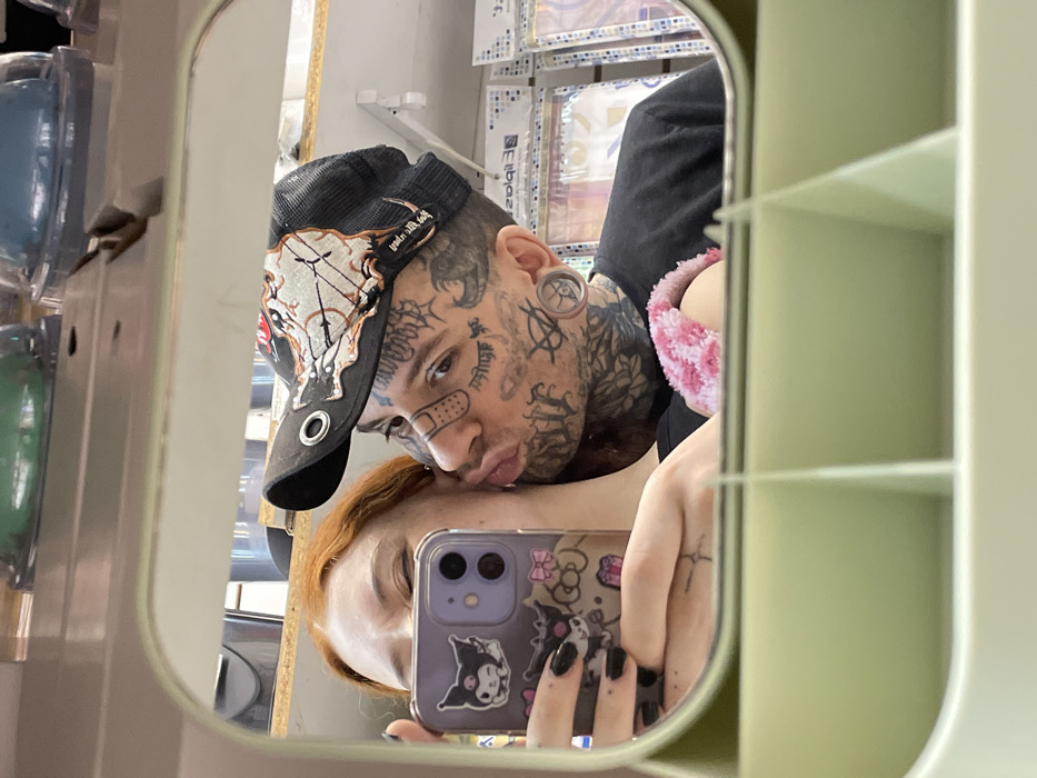 The image is a mirror selfie depicting two individuals in a retail store setting. A female subject with red hair and black fingernails is partially visible on the left, holding a purple iPhone adorned with Sanrio character stickers, capturing the reflection. Her face is turned towards the male subject. The male subject, on the right, has extensive tattoos covering his face and neck, including a stylized band-aid on his right cheek and large ear plugs. He wears a black and beige trucker-style baseball cap with a detailed pattern and a black t-shirt. His lips are pursed in a kissing gesture directed towards the female's cheek. A pink, fuzzy strap or accessory is visible on his right shoulder. The background, blurred and reflected in the mirror, shows shelves stocked with various retail products, including items in blister packs. While the scene takes place in a store, there are no specific identifying visual markers to pinpoint a precise location within Buenos Aires, Argentina.