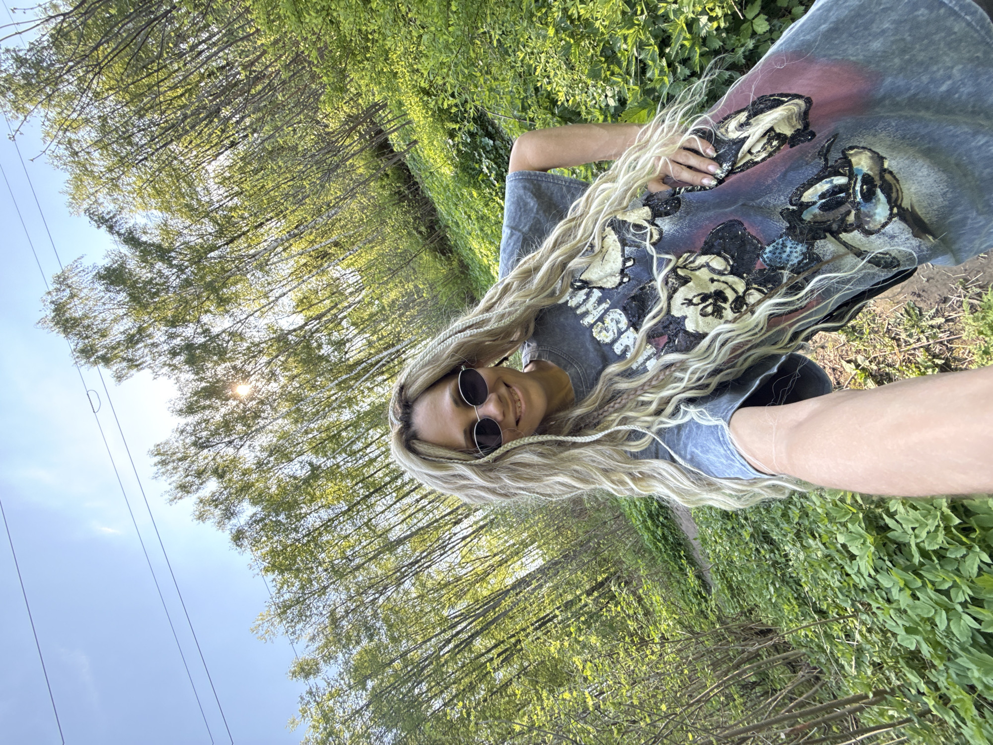 A woman stands outside in a wooded area, holding the camera to take a selfie. She is wearing a t-shirt, sunglasses, and has long blonde hair with braids. The environment includes trees, green foliage, and a sky with some visible sunlight. The location is in Rusanovo, Russia.