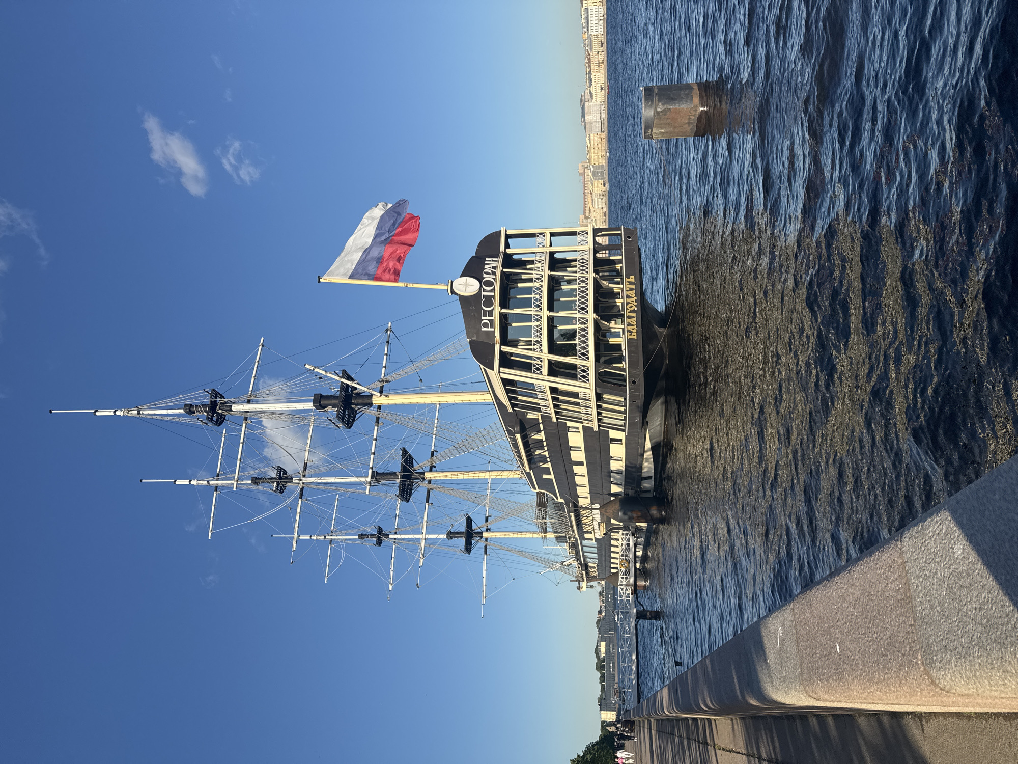 The image shows a large, three-masted sailing ship docked on the Neva River in Saint Petersburg, Russia. The ship, named "РЕСТОРАН" (Restaurant) and "БЛАГОДАТЬ" (Grace), is painted black and white and has multiple decks with numerous windows. A Russian flag flies from the stern. The ship is moored alongside a granite embankment, where a paved walkway is visible. Several people are walking on the embankment. The background reveals the city skyline of Saint Petersburg, with historic buildings and a clear blue sky with a few wispy clouds. The water of the Neva River is dark blue with ripples. A cylindrical, dark metal buoy is visible in the foreground, to the right of the ship. The lighting suggests it is a sunny day, likely in the late afternoon or early evening, given the long shadows cast on the embankment.