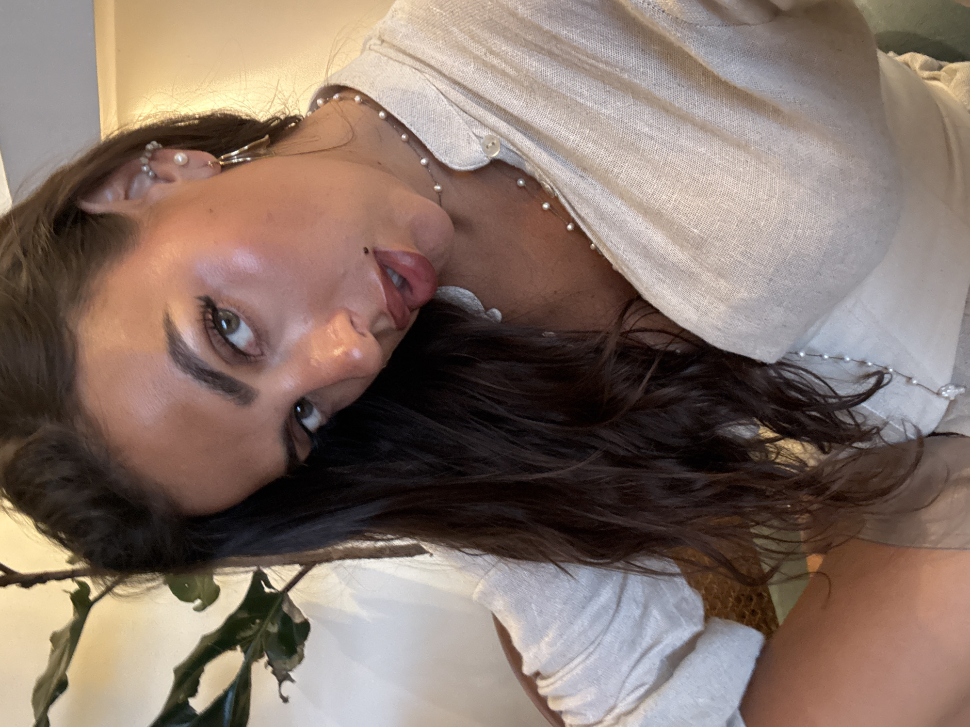 A woman with brown hair and fair skin is shown from a close, tilted perspective. She is wearing a light-colored shirt with buttons and a pearl necklace, and has several piercings in her left ear. Her eyes are open, and her lips are slightly parted. The background includes a plant with green leaves. The location of North Kuta, Indonesia is not apparent from the image itself, as there are no clear landmarks or contextual clues that would confirm this location.