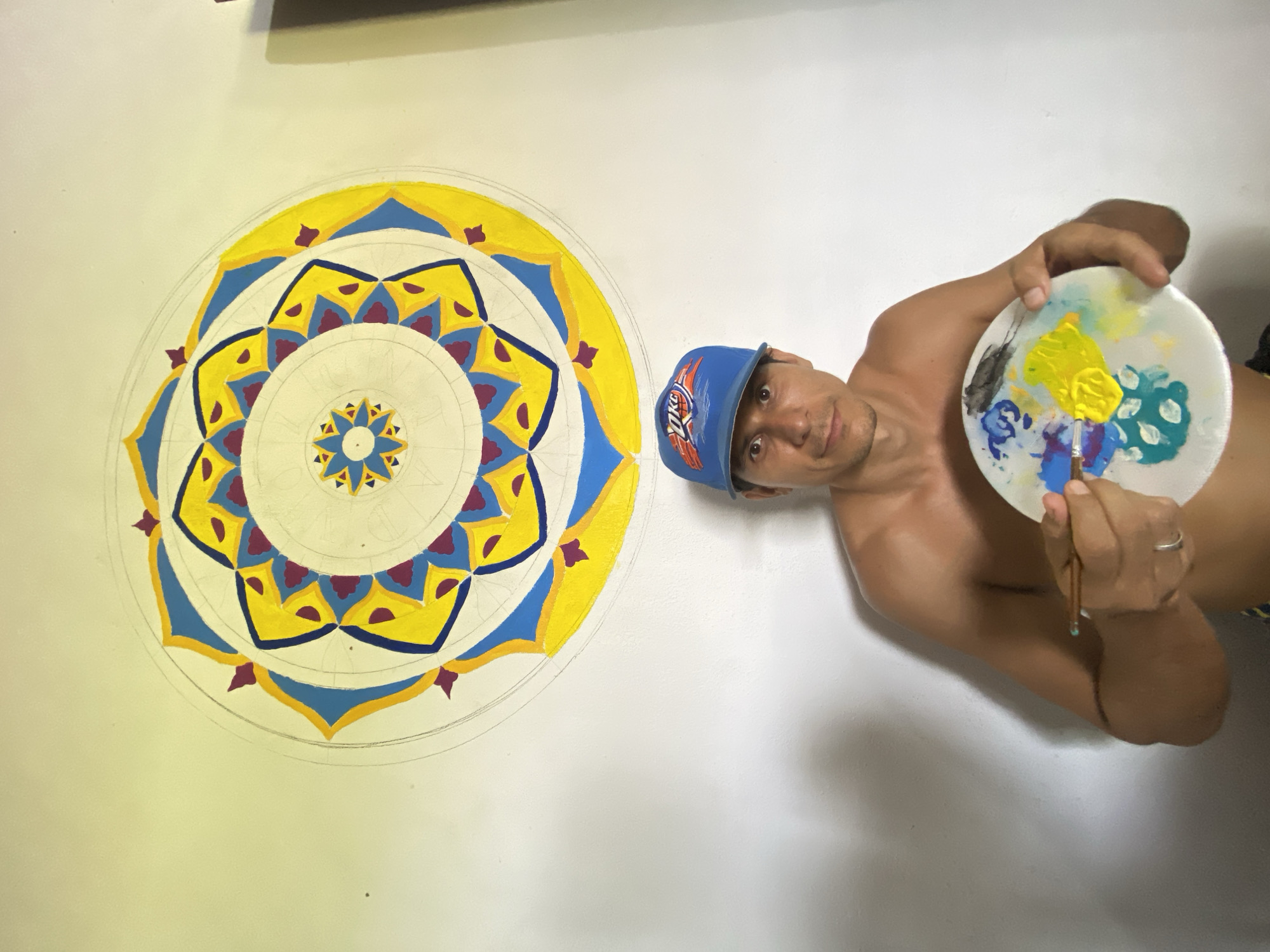 A man with light skin, wearing a blue baseball cap with an "OKC" logo, is holding a white paint palette with various colors of paint (yellow, blue, green, black) and a paintbrush. He's shirtless and wears a wedding ring on his left ring finger. The man is positioned next to a large, colorful mandala painting on a white wall. The mandala features concentric circles and repeating patterns in yellow, blue, and purple. Sketch lines are still visible in unpainted areas of the design. The location is indicated as Los Velásquez, Venezuela.