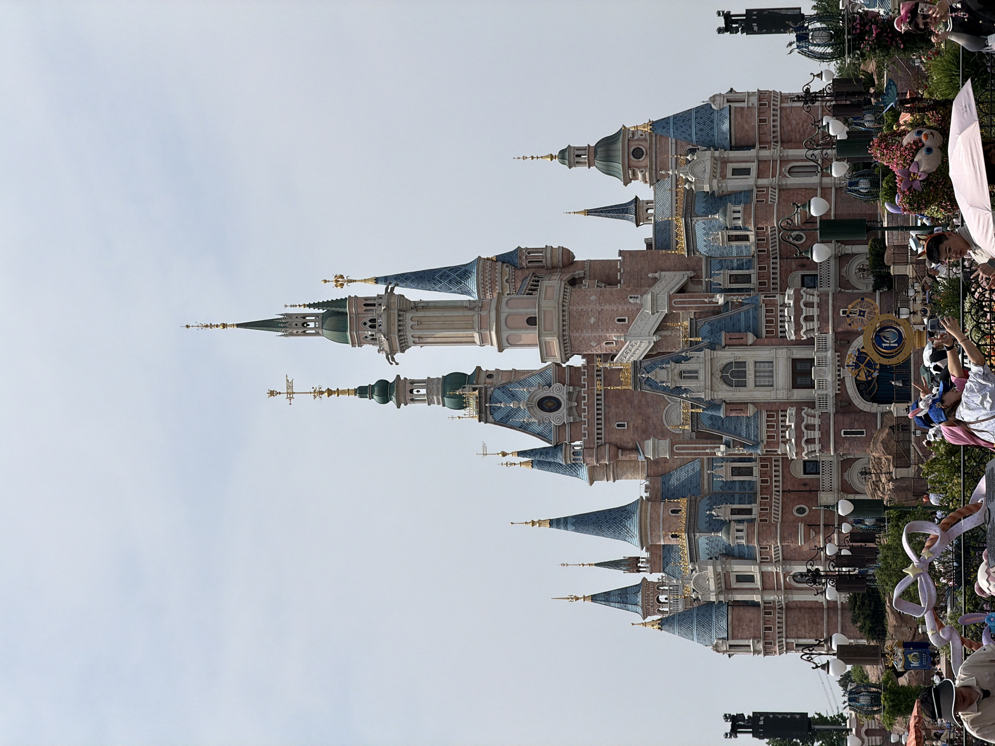 The image shows a view of the Shanghai Disneyland castle in Pudong, China. The castle features various towers and spires with blue roofs and pink walls. People are visible along the perimeter, some taking pictures and some holding balloons. There are trees and other landscaping features in the foreground. Street lamps are also visible. The sky is overcast.