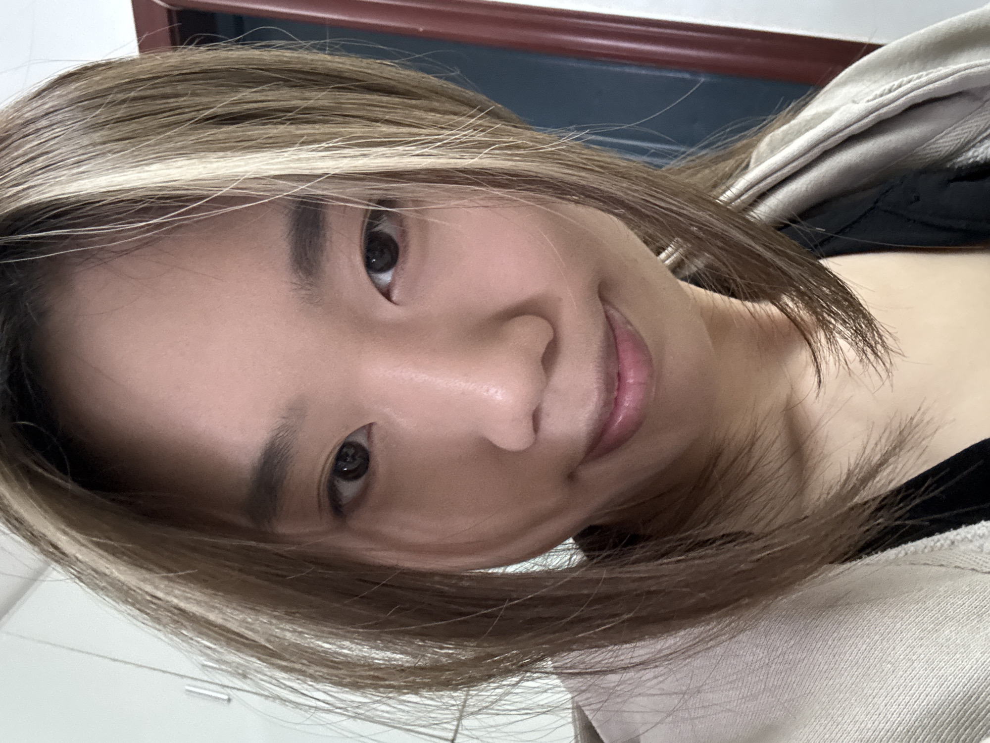 A close-up, angled selfie of a woman with light brown hair and blonde highlights. She has dark eyes and is partially smiling. She is wearing a black top underneath a light-colored jacket or hoodie. The background is indistinct, showing parts of a room with a dark blue wall trim. There is no information to determine if this image was taken in Shapingba District, China.
