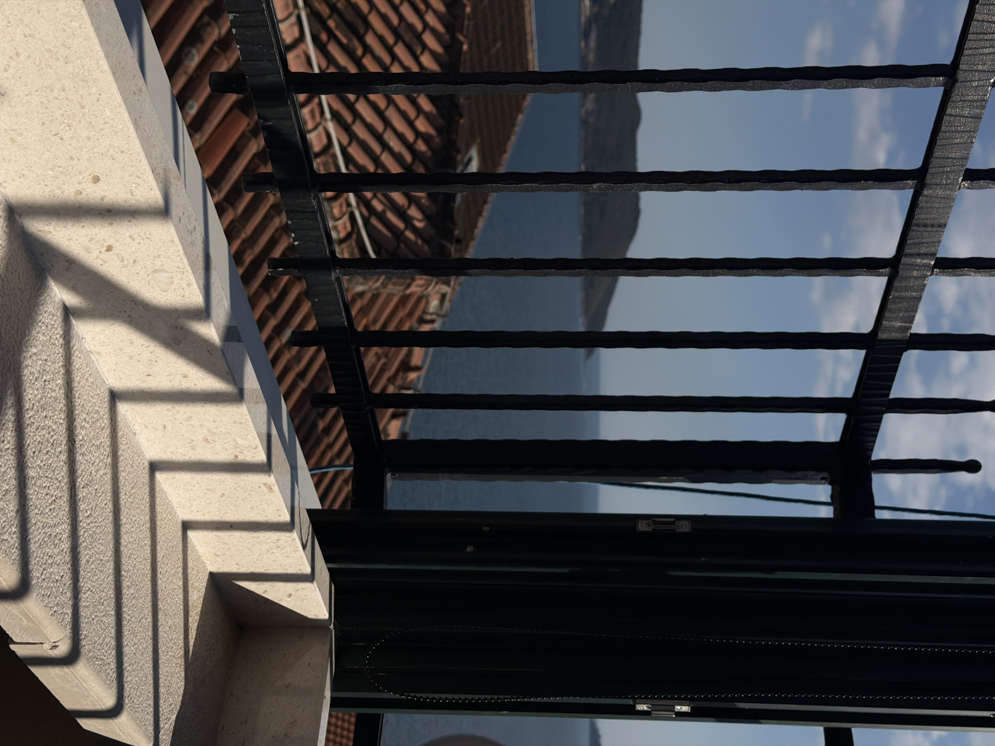 The image shows an outdoor scene, possibly in Trebesin, Montenegro, due to the presence of terracotta roof tiles. A stone balustrade casts shadows, next to a black metal railing, through which a body of water and a mountainous landscape are visible. Parts of a building are also visible through the railing. A black roller shade is partially down with an attached cord. The sky, with some clouds, is reflected in the panes of glass in the shade.