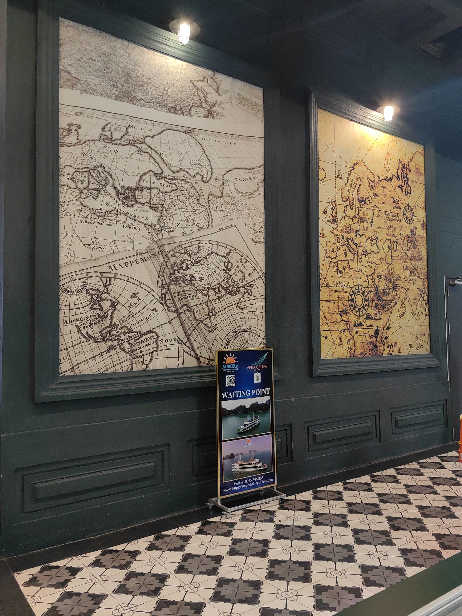 The image depicts an interior wall with two large, framed antique world maps. The first map on the left, primarily in shades of brown and beige, appears to show Europe, Africa, and parts of the Americas. Text and geographical features are visible. The second map on the right, rendered in similar colors but with a yellower tone, seems to focus on Europe, Africa, and Asia. Below the first map sits a small, advertising sign displaying the words "WAITING POINT" and some pictures of what appears to be a cruise ship with the words "AURORA VERA CRUISE". The wall is painted in a dark teal or green color with raised paneling. The floor is covered in a patterned tile of alternating black-and-white geometric shapes. The maps are illuminated by overhead lighting. The geographical location is implied, due to the sign, to be somewhere with access to "Ha Long Bay". The city is named as Bai Chay Ward, Vietnam.