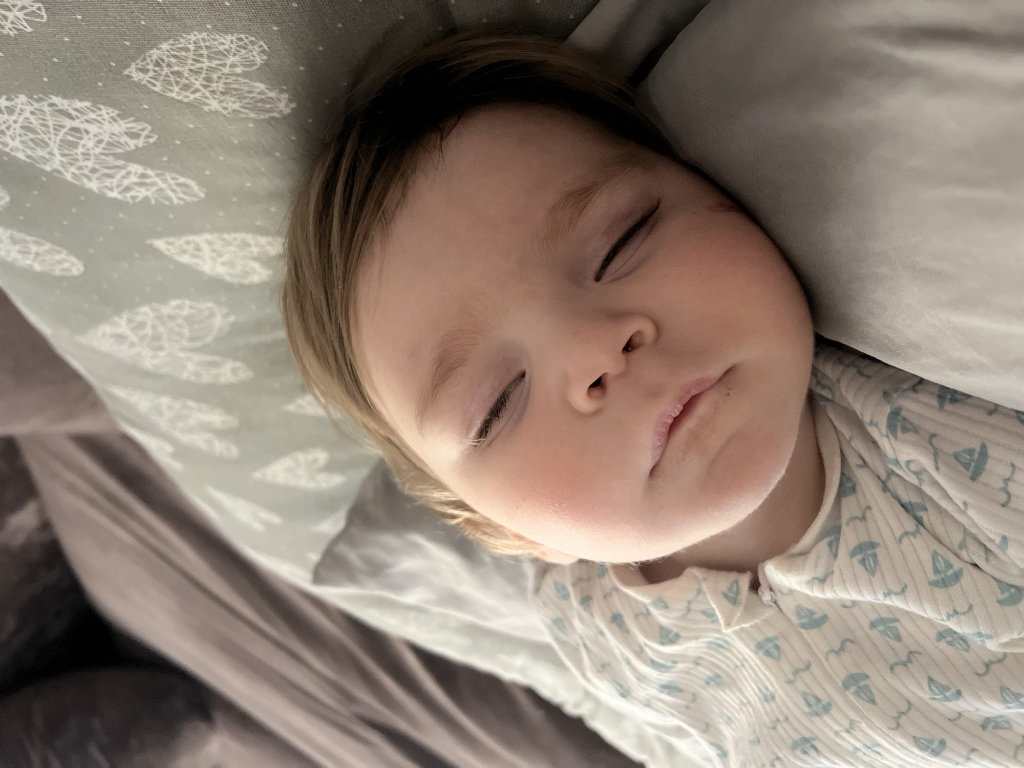 A baby is sleeping on a pillow. The pillowcase is light gray with a pattern of white shapes. The baby is wearing a white onesie with blue sailboat and bird designs. The background is out of focus, but appears to be bedding. The location is not discernible from the image.