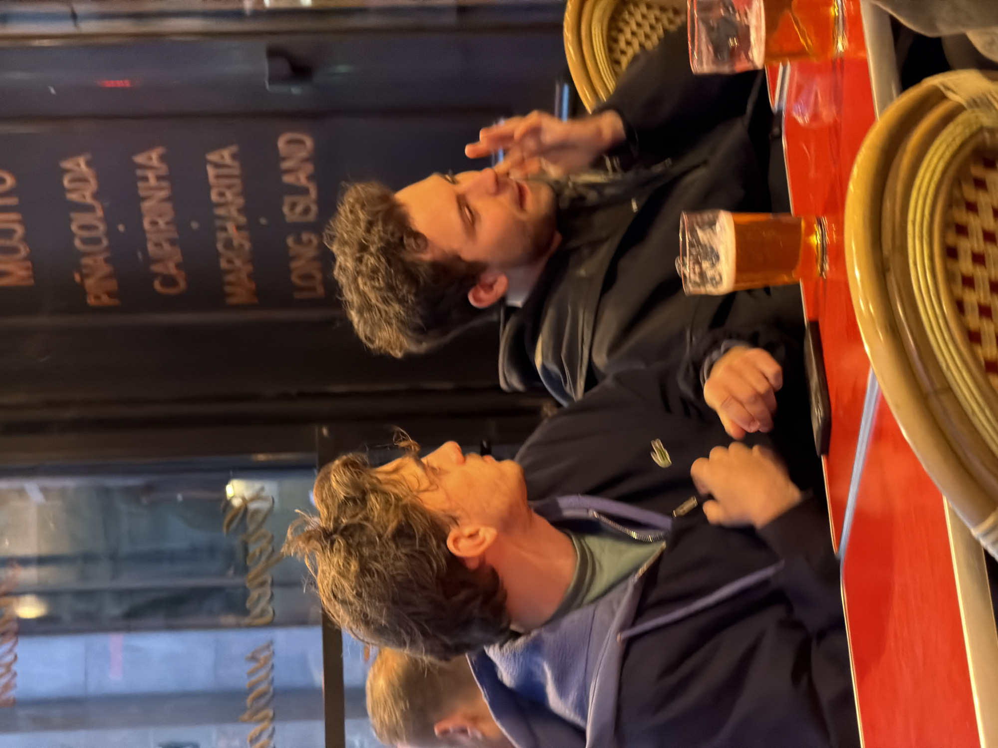 Two men are seated at an outdoor cafe in Paris, France. The cafe menu lists alcoholic drinks, including mojitos, pina coladas, capirinhas, margaritas, and long island iced teas. Both men are wearing jackets and appear to be engaged in conversation, with the first man gesturing with his hand. There is a pint of beer, and another drink on the red tabletop. They are sitting in classic Parisian wicker chairs. The scene is taken at an angle.