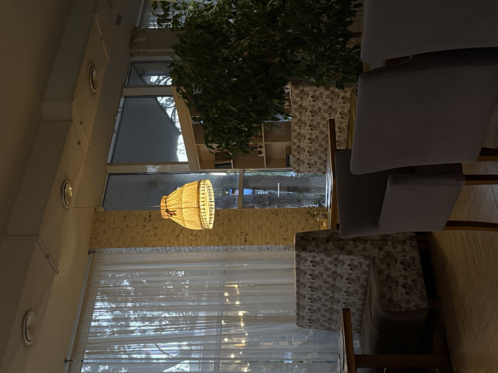 The image depicts the interior of a cafe, likely located in Abinsk, Russia, during what appears to be late afternoon or early evening, given the ambient lighting and the light filtering through the windows. The foreground features two dark gray upholstered armchairs facing towards the viewer and a table with a wooden frame. To the left, a wooden table is partially visible with a placemat.
Behind the armchairs, a floral-patterned banquette seating area is prominent, with one section extending to the left and another to the right, partially obscured by a large potted plant. The banquette is situated near a window with sheer white curtains.
The walls are a light-colored brick. A distinctive yellow lampshade with a fringed edge hangs from the ceiling. Large windows reveal trees and the exterior environment. Behind the banquette and plant, a wooden bookshelf with various items is visible. The flooring is light brown parquet.
No people are visible in the image. The scene conveys a sense of quiet ambiance within the cafe.