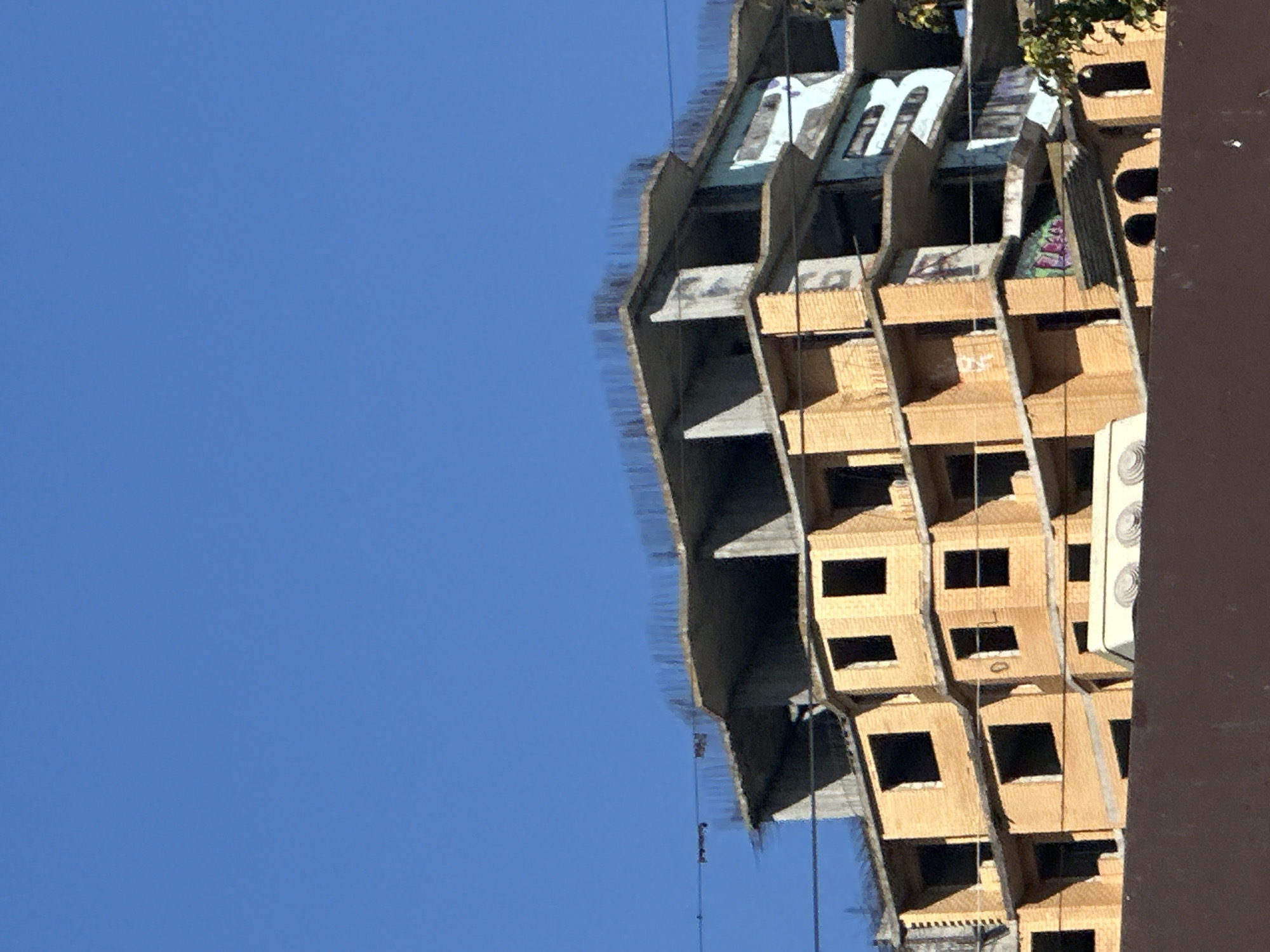 The image shows a multi-story building under construction against a clear blue sky. The building appears to be made of brick and concrete. Some floors are partially constructed, with exposed rebar on the top levels. There are visible windows that are dark, suggesting they are either not yet installed or are covered. Graffiti is visible on some sections of the building, particularly on the lighter colored exterior walls. The lower part of the image is obscured by a dark, horizontal structure, possibly a fence or another building. The scene suggests an urban environment, and the presence of unfinished construction indicates it is likely a new development.
