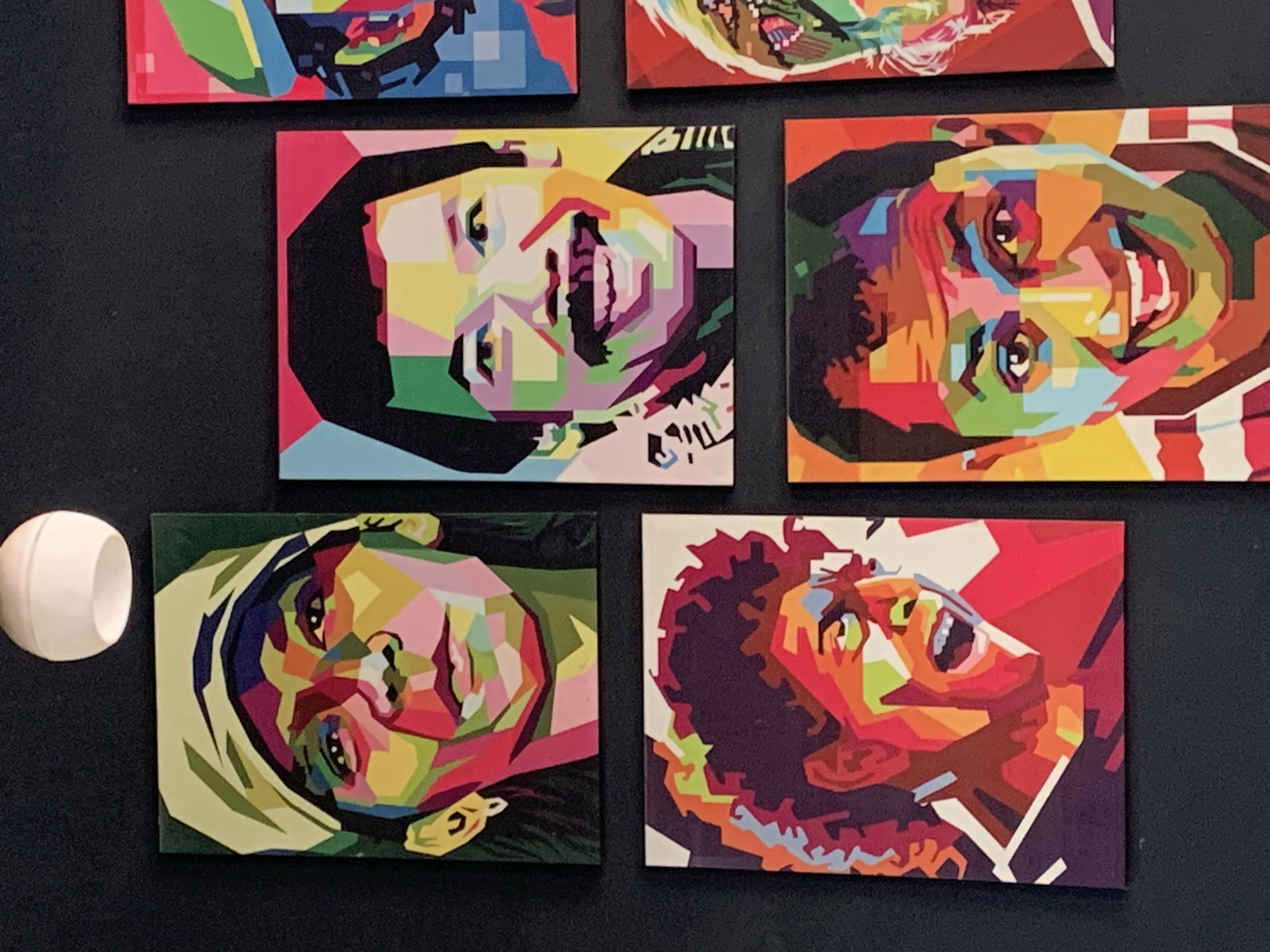 The image shows a wall adorned with several vibrant, geometric portraits of people. The artworks are rendered in a pop art style, utilizing bold, flat colors and sharp angles to depict facial features.
Specifically, the arrangement includes:
* **Top Left:** A portrait of a person wearing a light-colored cap or bandana, with their face partially visible.
* **Top Right:** A partial view of another portrait, primarily showing a green cap and elements of a colorful face.
* **Middle Left:** A portrait of a person with curly hair, their mouth open in a shout or exclamation.
* **Middle Right:** A portrait of a man with a mustache, smiling broadly.
* **Bottom Left:** A close-up of a person's face, with their mouth wide open.
* **Bottom Right:** A portrait of a smiling person with a colorful patterned shirt.
These artworks are mounted on a dark, textured wall. A white, spherical light fixture is visible in the upper left corner of the frame. The overall impression is of a gallery or decorative display featuring stylized portraits.
The image is attributed to the city of Palohlada, Indonesia.