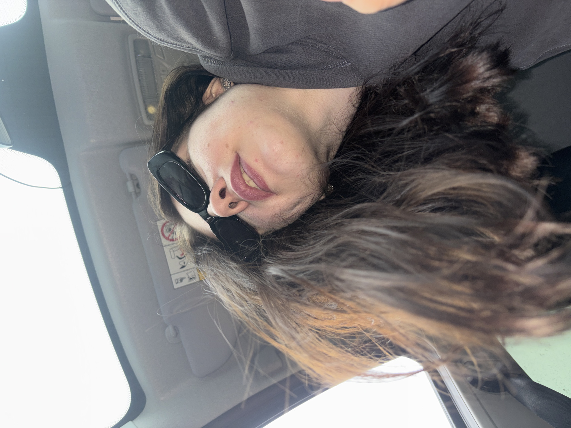 A woman is in a car, wearing sunglasses and a gray hooded sweatshirt. Her hair is dark and partially obscures the view. The car's interior is visible, including the sun visor with airbag warnings. The location cannot be determined from this image.