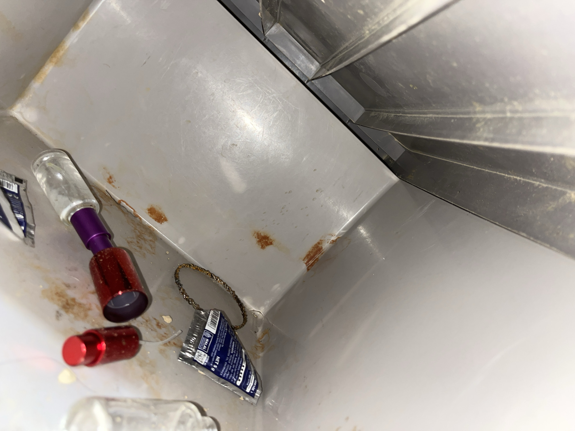 The image shows a close-up, high-angle view of the interior of a light-colored plastic bin, possibly a trash can, located indoors. The bin's surfaces show signs of wear, including scratches and reddish-brown stains, indicative of rust or dried liquid. Several items are present within the bin. On the left side, a clear glass bottle with a metallic purple and red nozzle is partially visible. Next to it, a red cap from a bottle lies on its side. In the center, a small, silver-colored packet with blue and white labeling, including text that appears to be in Indonesian and mentions "NET 5g," is attached to a thin, twisted metal chain. The packet also has a small barcode. Further to the right, the edge of a metallic structure, likely part of the bin's lid or an adjacent appliance, is visible. The lighting is dim, casting soft shadows. The overall impression is one of a discarded or unused item within a utilitarian container, possibly in a commercial or industrial setting given the metallic elements. There is no indication of the specific location within Kota Kuala Simpang, Indonesia.