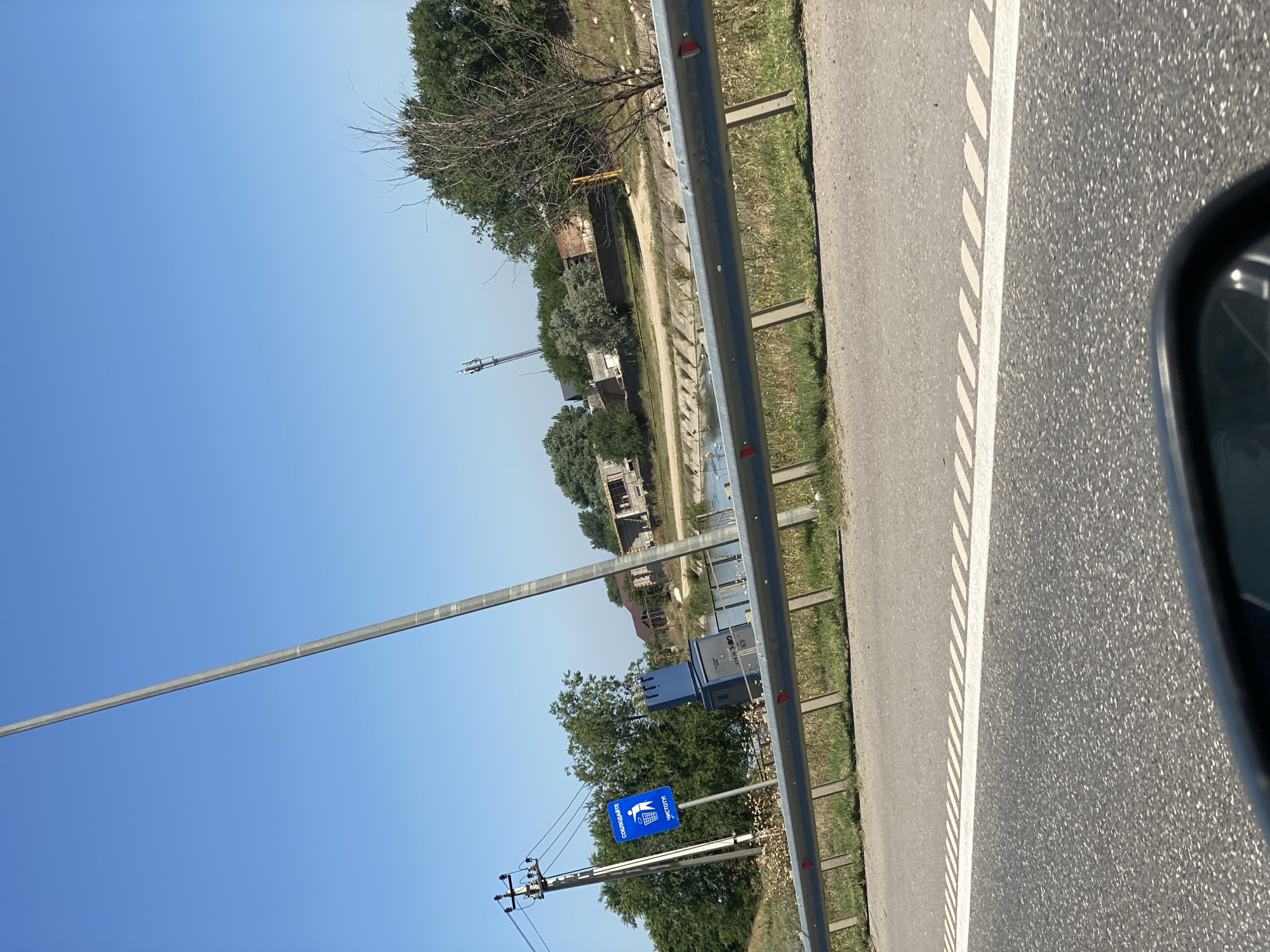 This is a medium-angle, eye-level, outdoor, daytime, natural light, slightly tilted photographic image of a roadside scene. The perspective is from a vehicle traveling on a paved road, indicated by the asphalt in the foreground and the white dashed lane divider. A metallic guardrail runs parallel to the road, separating it from a grassy embankment and a body of water with concrete edges.

On the left side of the frame, a utility pole with multiple wires is visible. Adjacent to it, a blue sign with white text and an image of a trash can with an 'X' over it is posted, likely indicating a no-littering rule. A dark blue electrical cabinet with vents is situated behind the sign, with some Cyrillic lettering and the number "63" visible on it.

Further back, a body of water runs alongside the road, suggesting a canal or river. On the opposite bank of the water, there are several houses under construction and some completed buildings with brick and concrete structures. Trees and vegetation are scattered throughout the background. A tall communications tower with antennas is also visible in the distance. The sky is clear and blue. The overall scene depicts a rural or semi-rural environment, possibly in the city of Stalskoye, Russia, characterized by infrastructure and residential development alongside a waterway.