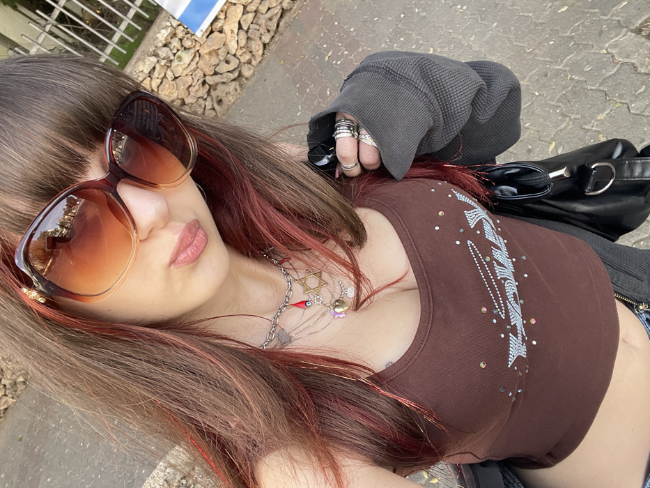 A young individual is depicted in an outdoor, upward-angle selfie-style photograph. The subject has long brown hair with reddish streaks and bangs, and is wearing large brown-toned sunglasses with gradient lenses. Their lips are pursed. They wear a brown spaghetti-strap crop top featuring a light blue and multi-colored rhinestone design on the chest, a gray waffle-knit long-sleeved garment, and partially visible blue denim. Multiple layered necklaces adorn their neck, including a silver-colored chain with charms such as a red chili, a blue evil eye, and a Star of David, alongside thinner chains with other charms like a heart. The subject's right hand (viewer's left) is partially obscured by the sleeve and holds a dark object, possibly a black bag strap, and displays numerous rings on the fingers. A black shoulder bag with silver-colored hardware is visible. The background consists of a paved ground surface, with a fence, rocky terrain, and a partially visible blue and white object (consistent with Israeli public signage) on the right side. The scene is located in the city of Raanana, Israel.