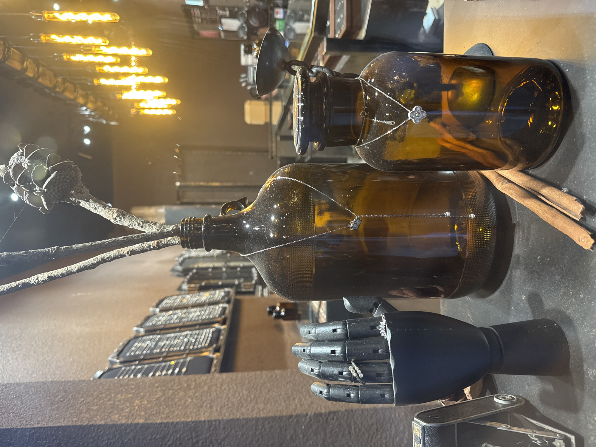 The image depicts a still life arrangement on a dark, textured surface. In the foreground, a black prosthetic hand, segmented and articulated, displays two rings on its fingers. To the left of the hand is a vintage camera. Behind the hand and camera are two large, amber-colored glass bottles. The larger bottle to the left has a branch with a dried, cone-like structure at the top inserted into its opening, and a silver necklace with small pearl-like pendants hangs from it. The smaller bottle to the right also has a silver necklace with a clover-shaped pendant hanging from its opening. Two cinnamon sticks lie diagonally in front of the bottles. In the background, the environment appears to be an indoor retail space, possibly a jewelry store, with display shelves filled with items and a row of illuminated Edison-style light bulbs hanging from the ceiling. The overall aesthetic is dimly lit with warm, ambient light. There are no people visible in the image, and no specific location in Hanoi, Vietnam, can be identified from the visual information provided.