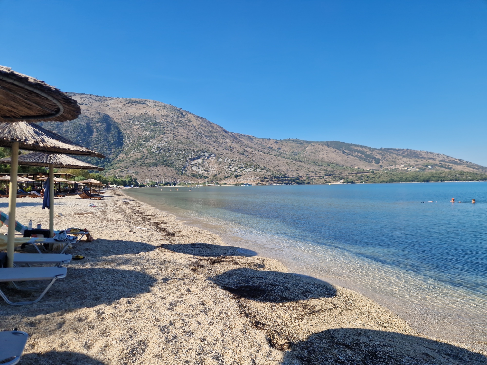 The image depicts a beach scene in Plataría, Greece, under a clear blue sky. In the foreground, several straw umbrellas cast shadows on the pebble-strewn beach. Beach chairs are visible, some occupied by individuals relaxing. The water is clear and calm, with a few swimmers visible in the distance. A gently sloping, tree-covered mountain range forms the backdrop. The overall atmosphere is peaceful and idyllic.