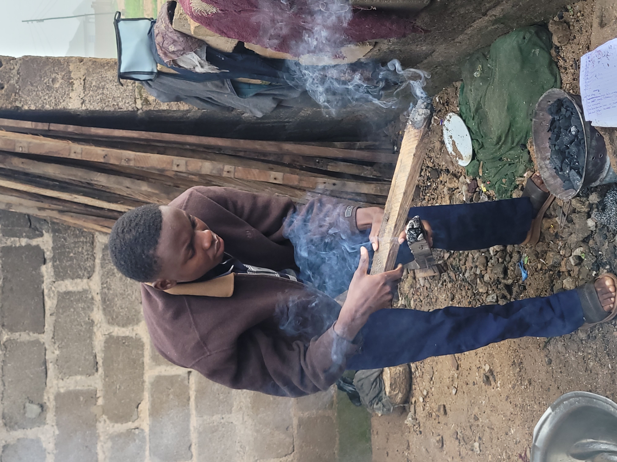 In Barakin Akawo, Nigeria, a young Black male is captured in a medium shot outdoors, holding a piece of wood that is smoldering with smoke. He wears a brown jacket with a tan collar over a darker shirt and dark blue pants. His feet are clad in dark sandals. The background reveals a rough brick wall, stacked wooden planks, and various items including a light blue bag, a red patterned cloth, and a green cloth covering something on the ground. A metal bowl filled with charcoal sits in the foreground, alongside a white plate, scattered debris, and a piece of paper with writing. The ground is covered in dirt and stones. The scene suggests an outdoor activity involving fire or smoke, possibly related to cooking or crafting.