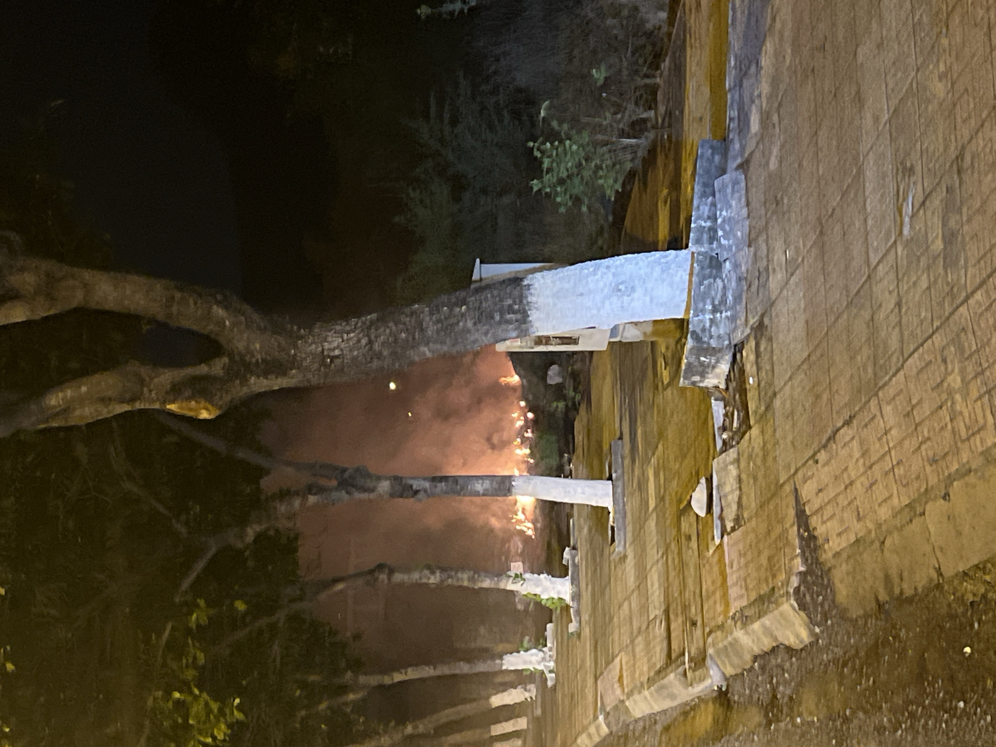 The image shows a nighttime scene in Bac Nha Trang Ward, Vietnam. A row of trees with whitewashed trunks lines a pedestrian walkway paved with rectangular tiles. The trees are partially obscured by a hazy orange glow, possibly from lighting or distant fires. The walkway is wet, reflecting the light. To the right, a building or structure borders the walkway. The overall impression is of an illuminated urban landscape at night.