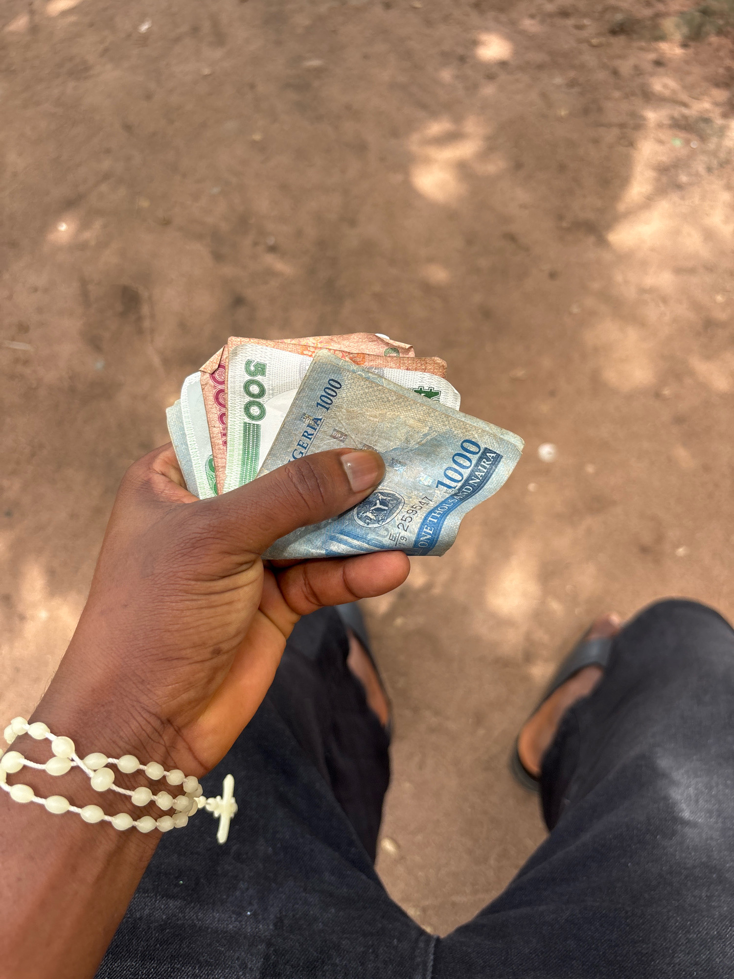 In Ihiagwa, Nigeria, a close-up shot shows a dark-skinned individual holding a fan of Nigerian Naira banknotes. The denominations visible are 1000 Naira, 500 Naira, and possibly others. The banknotes are neatly fanned out in the person's hand. The individual is wearing a pearl-like beaded bracelet with a cross charm on their left wrist. They are also wearing dark trousers and dark sandals. The background is a dusty, brownish ground, suggesting an outdoor environment in a rural or semi-urban setting. The lighting indicates it is daytime.