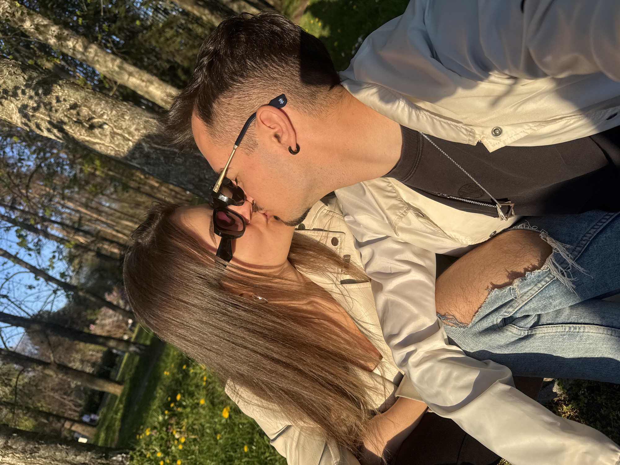 The image captures a couple kissing outdoors. The man has dark hair, sunglasses perched on his head, an earring in his left ear, and a silver necklace. He is wearing a black t-shirt and a light-colored jacket. The woman has long, brown hair and sunglasses. She is also wearing a light-colored jacket and ripped blue jeans. They appear to be sitting in a park or grassy area, with trees visible in the background. The scene is bathed in sunlight, suggesting it is daytime. The location appears to be in the city of Sofia, Bulgaria, based on the landscape and architecture that is often seen in this city.